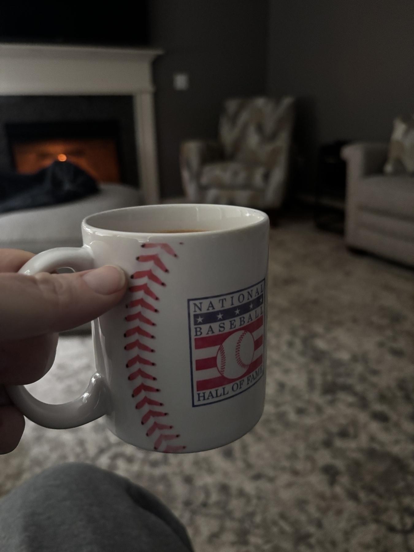 A person is holding a mug with a "National Baseball Hall of Fame" design in a cozy living room setting.