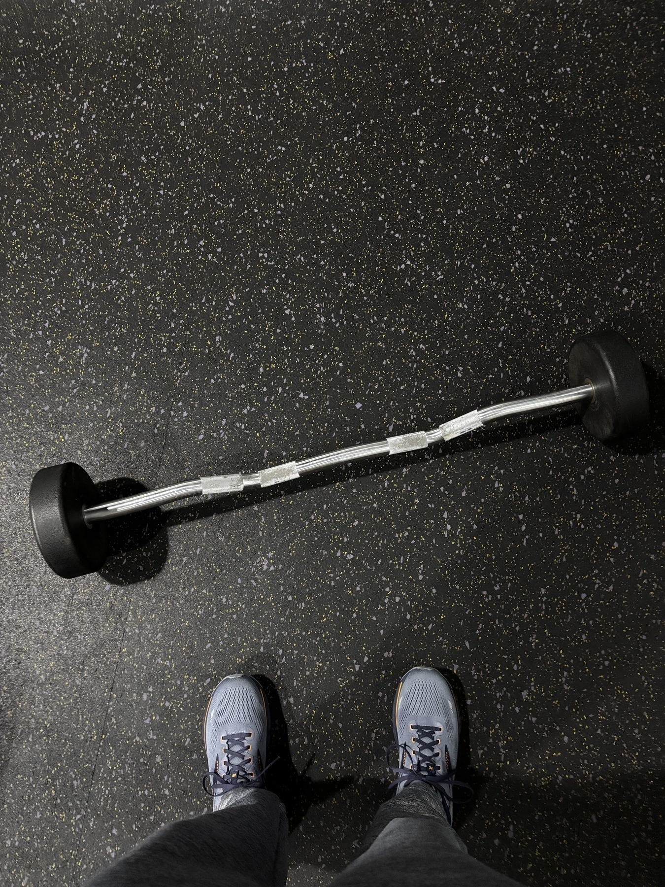 A barbell with attached weights rests on a speckled gym floor, viewed from above with a person's feet in athletic shoes visible at the bottom.