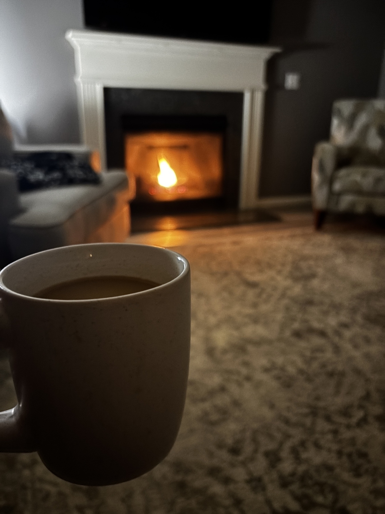 A cozy living room features a lit fireplace, a mug of coffee in the foreground, and seating in soft lighting.