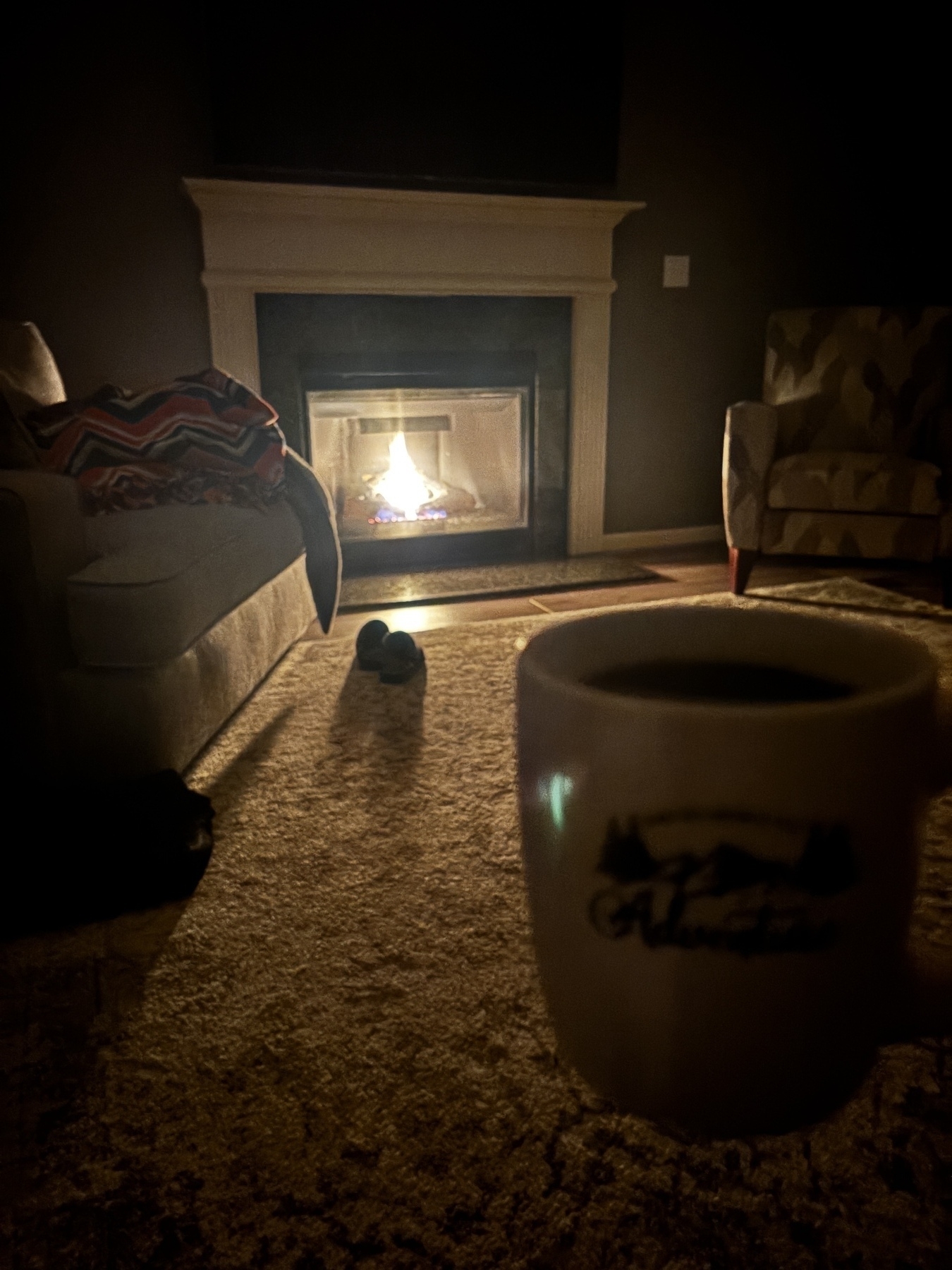 A cozy living room features a lit fireplace, a sofa with a colorful blanket, an armchair, and a mug in the foreground.