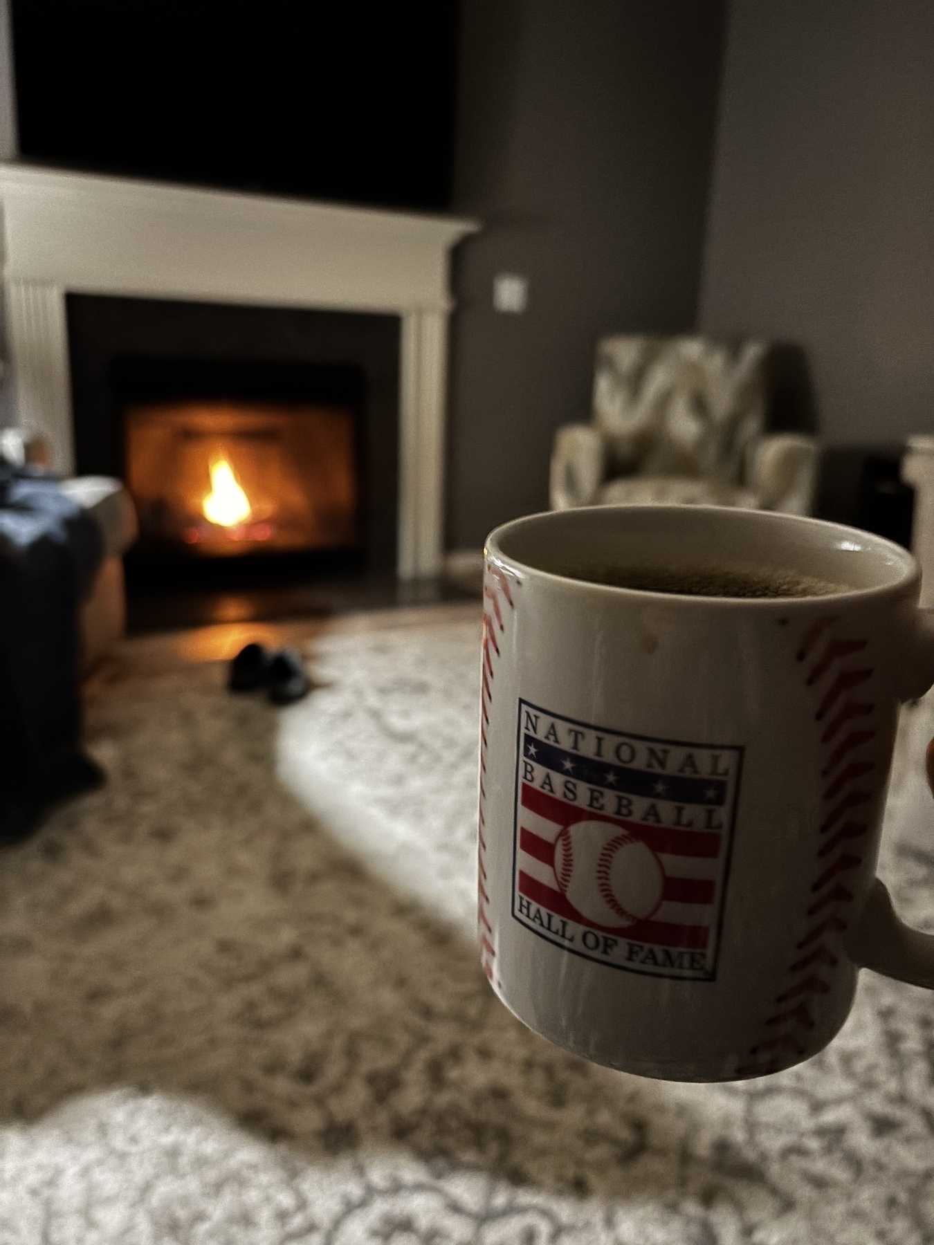 A cozy living room features a warm fireplace, a comfortable chair, and a mug with a National Baseball Hall of Fame logo.