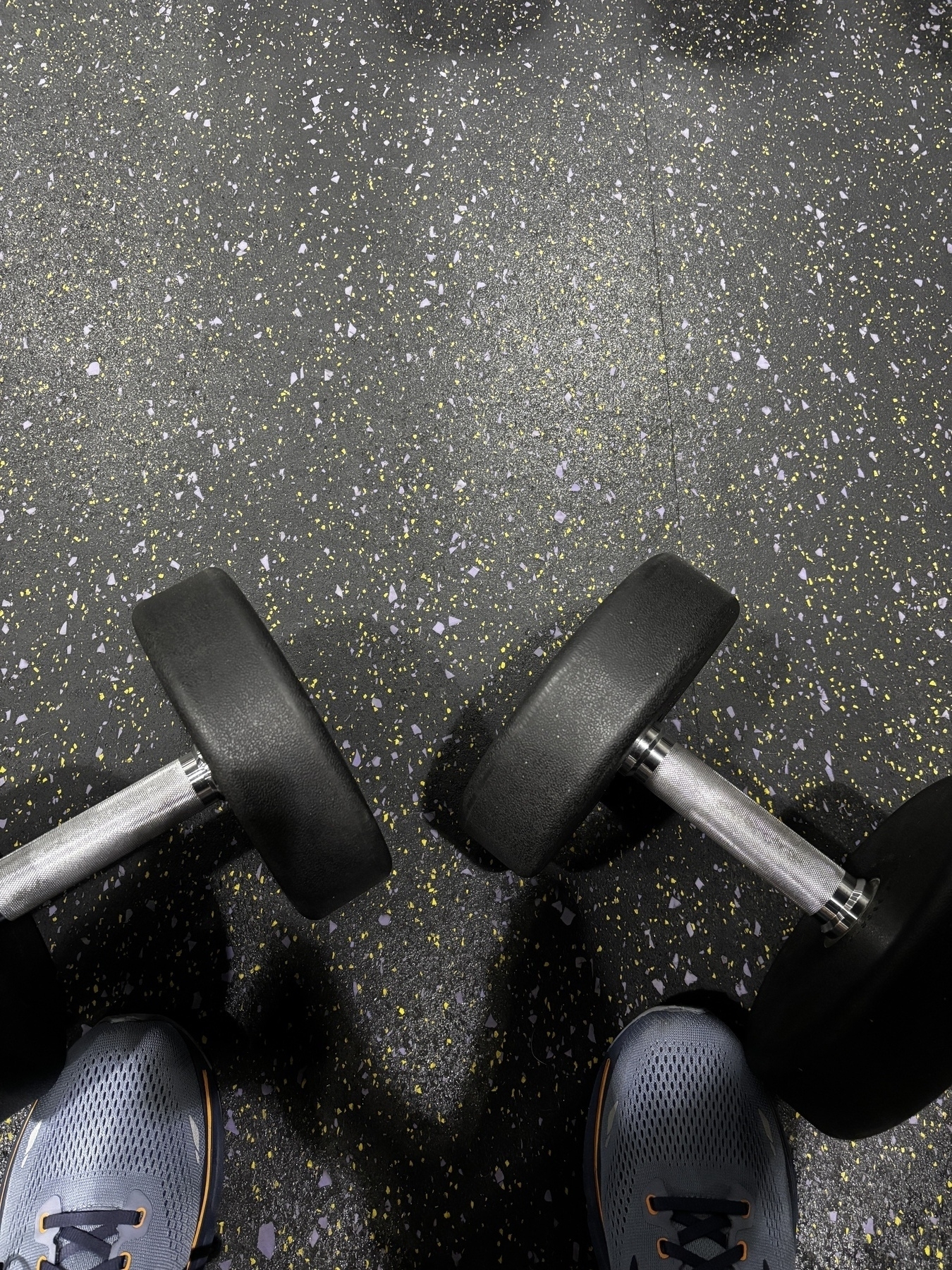 A pair of dumbbells is placed on a speckled gym floor next to someone's feet wearing sneakers.