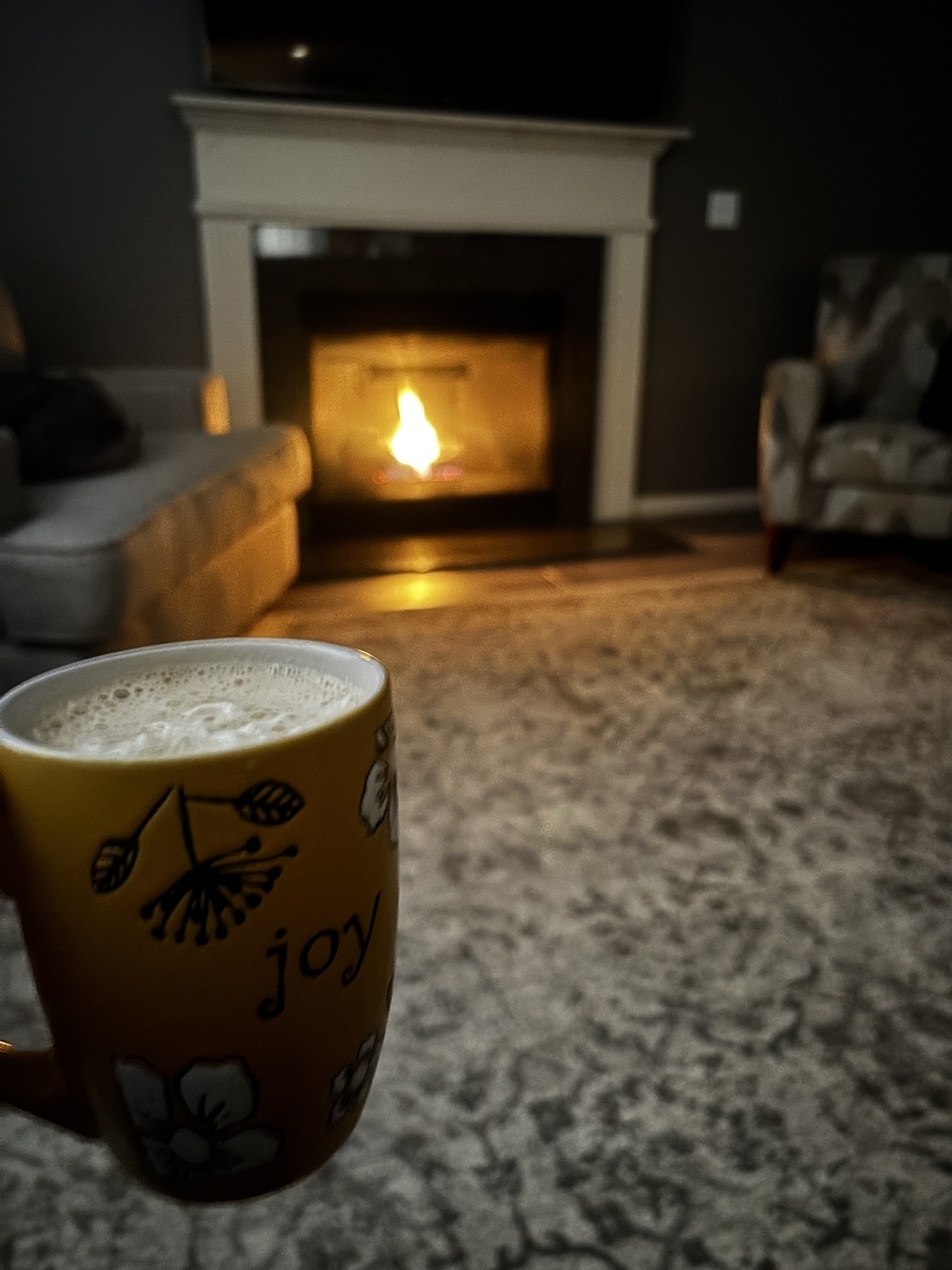 A cozy living room scene shows a person holding a yellow mug with a floral design and "joy" written on it, in front of a fireplace with a warm fire burning.