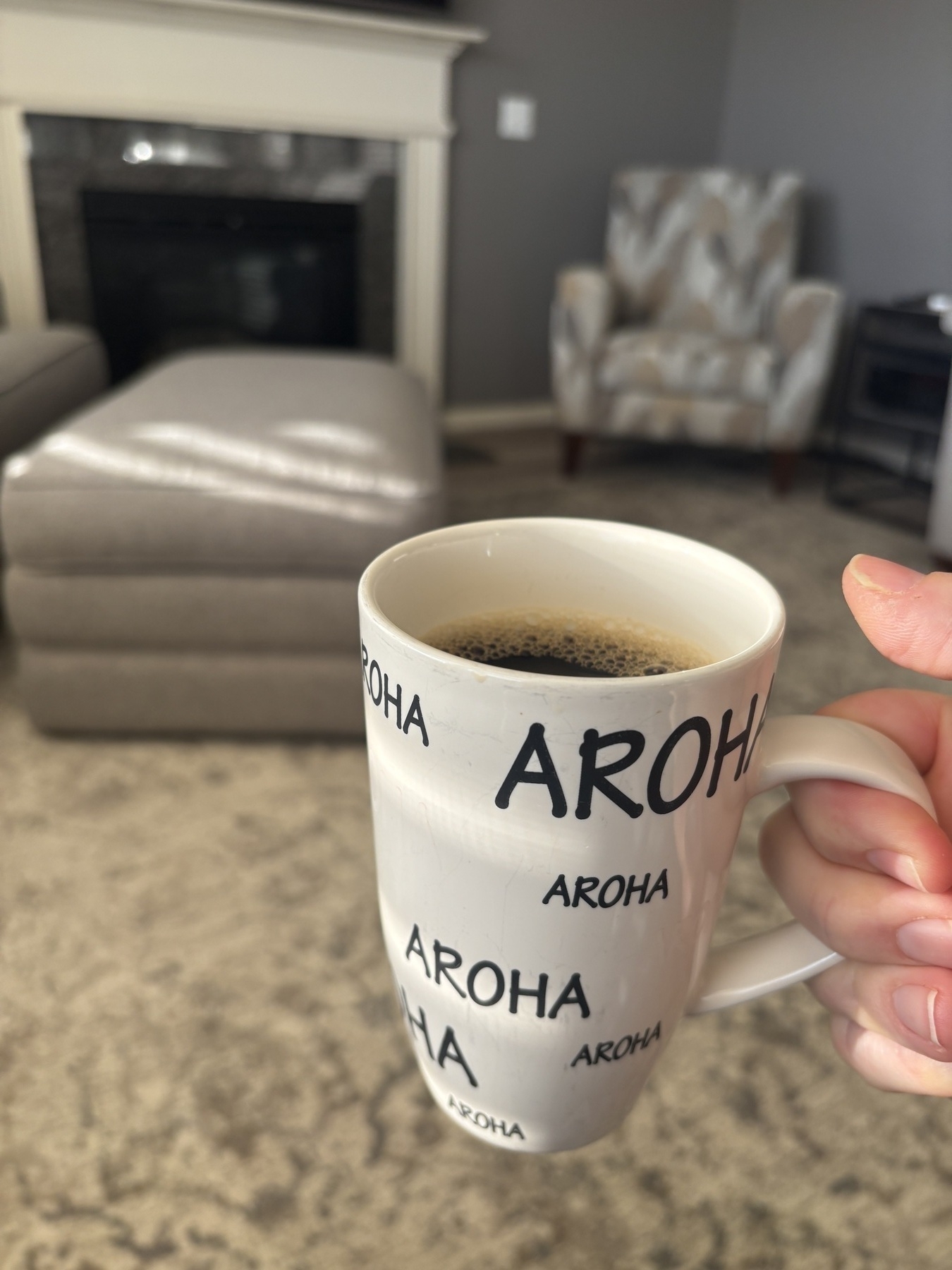 A cup of coffee with the word, “Aroha”, decorating it. This is Māori word for “love.” It is being held up in a cozy living room with the sun shining in. 