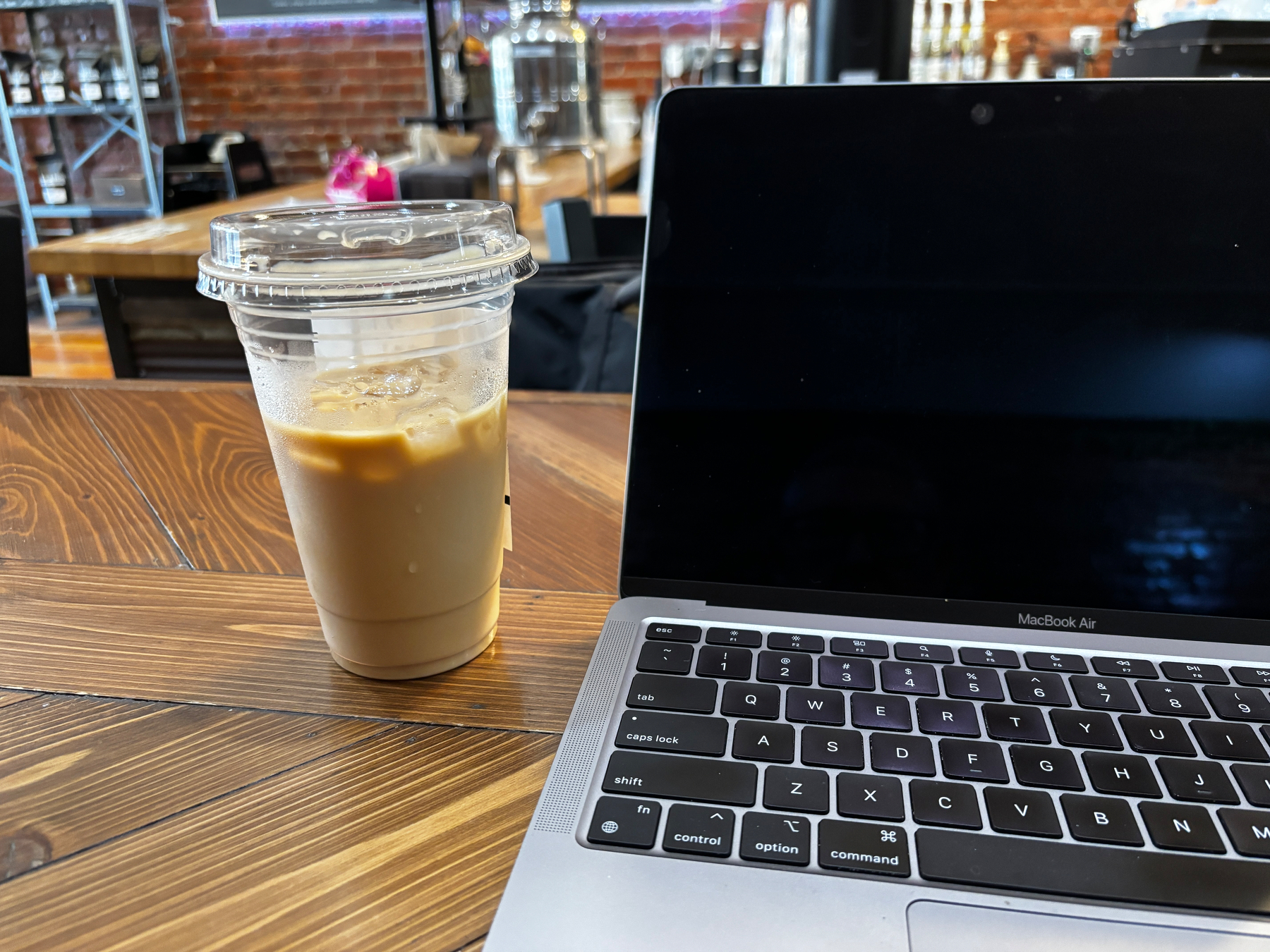 A laptop and a plastic cup of iced coffee are placed on a wooden table in a modern café setting.