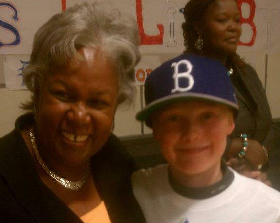 Sharon Robinson (L) and my son Ethan (R) wearing a Brooklyn Dodgers cap are smiling together indoors, with a woman in the background and a sign partially visible on the wall.