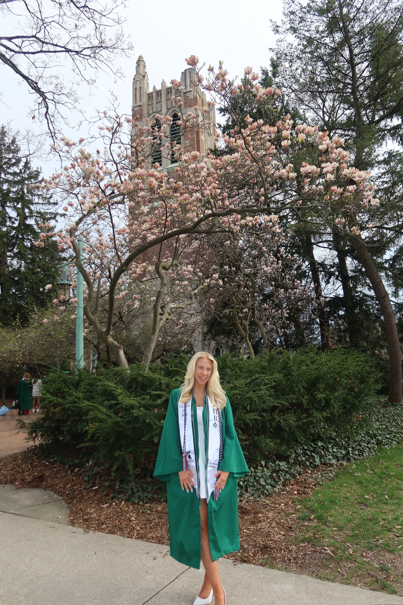 Libby in a green graduation gown stands in front of blooming trees with a large, ornate building in the background.