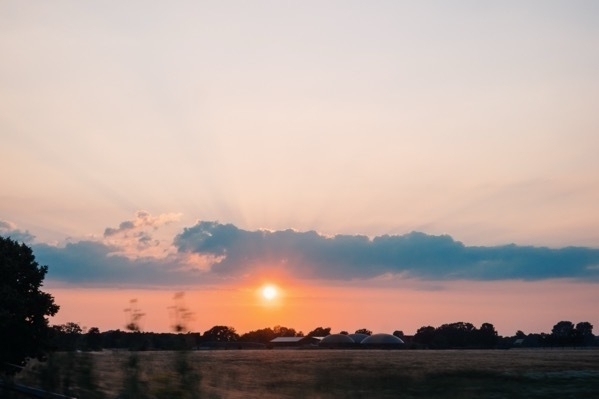 Ein Sonnenuntergang, über Feldern und ein paar Häusern. Der Himmel ist orange-gelb-blau gefärbt.