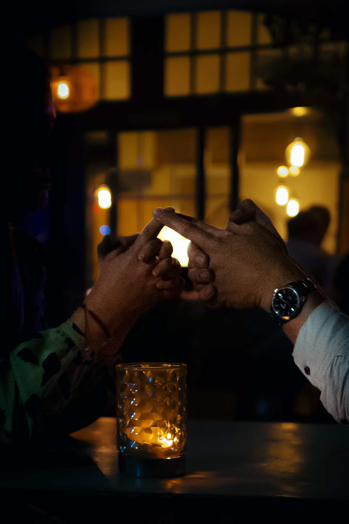 Zwei Hände sind ineinandergreifende Finger in einer dunklen, beleuchteten Umgebung, mit einem warmen Leuchten von einer Kerze in einem Glashalter auf dem Tisch. Der Hintergrund verfügt über sanft verschwommene Lichter, die eine gemütliche Atmosphäre schaffen.