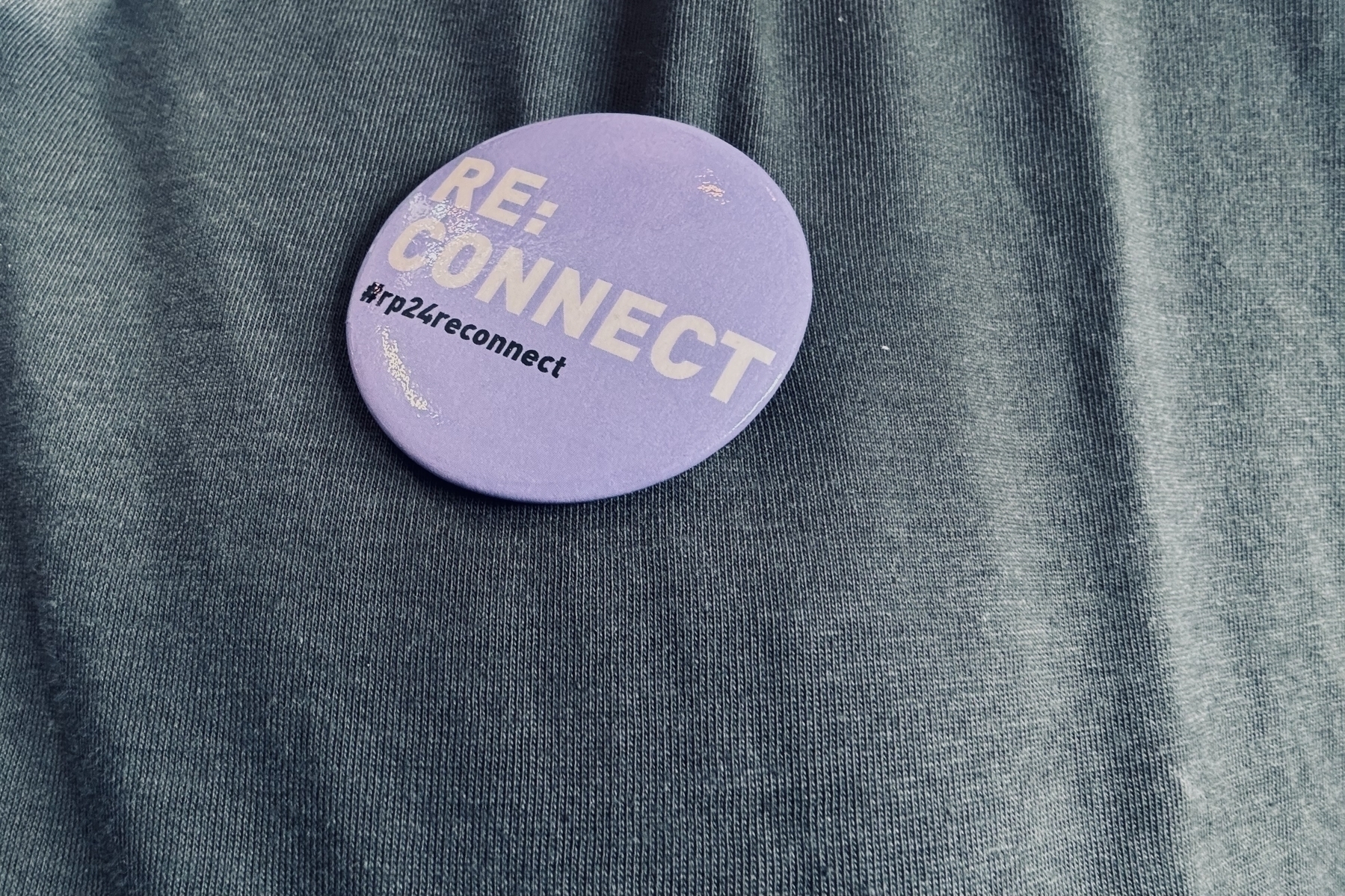 Ein lila kreisförmiger Button mit dem Text "RE:CONNECT #rp24reconnect", der an einen dunklen Stoff gepinnt ist.