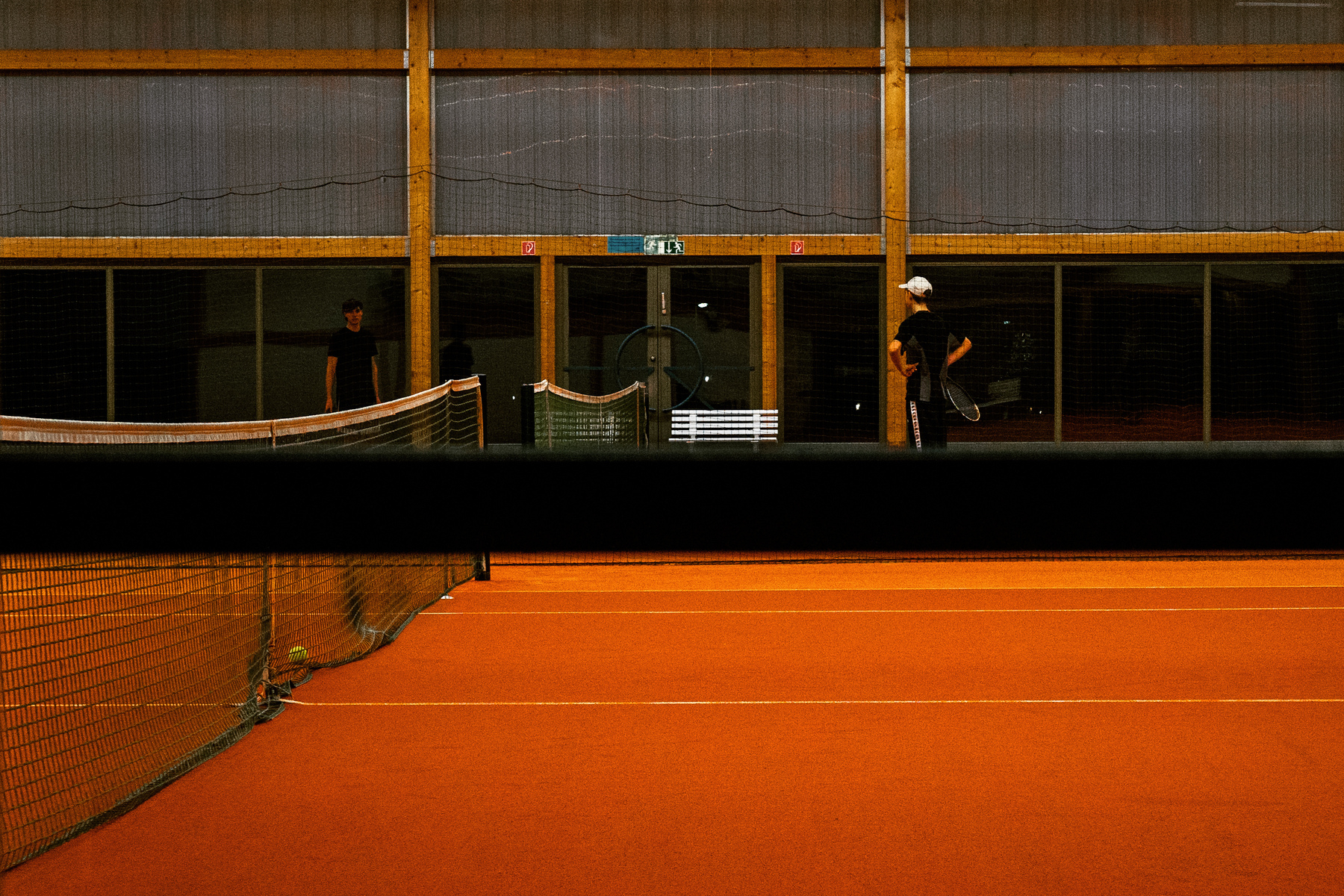 Ein Indoor-Tennisplatz mit zwei Spielern, die in der Nähe des Netzes stehen. Der Platz hat eine orangefarbene Oberfläche, und die Beleuchtung ist gedimmt, was eine stimmungsvolle Atmosphäre schafft.