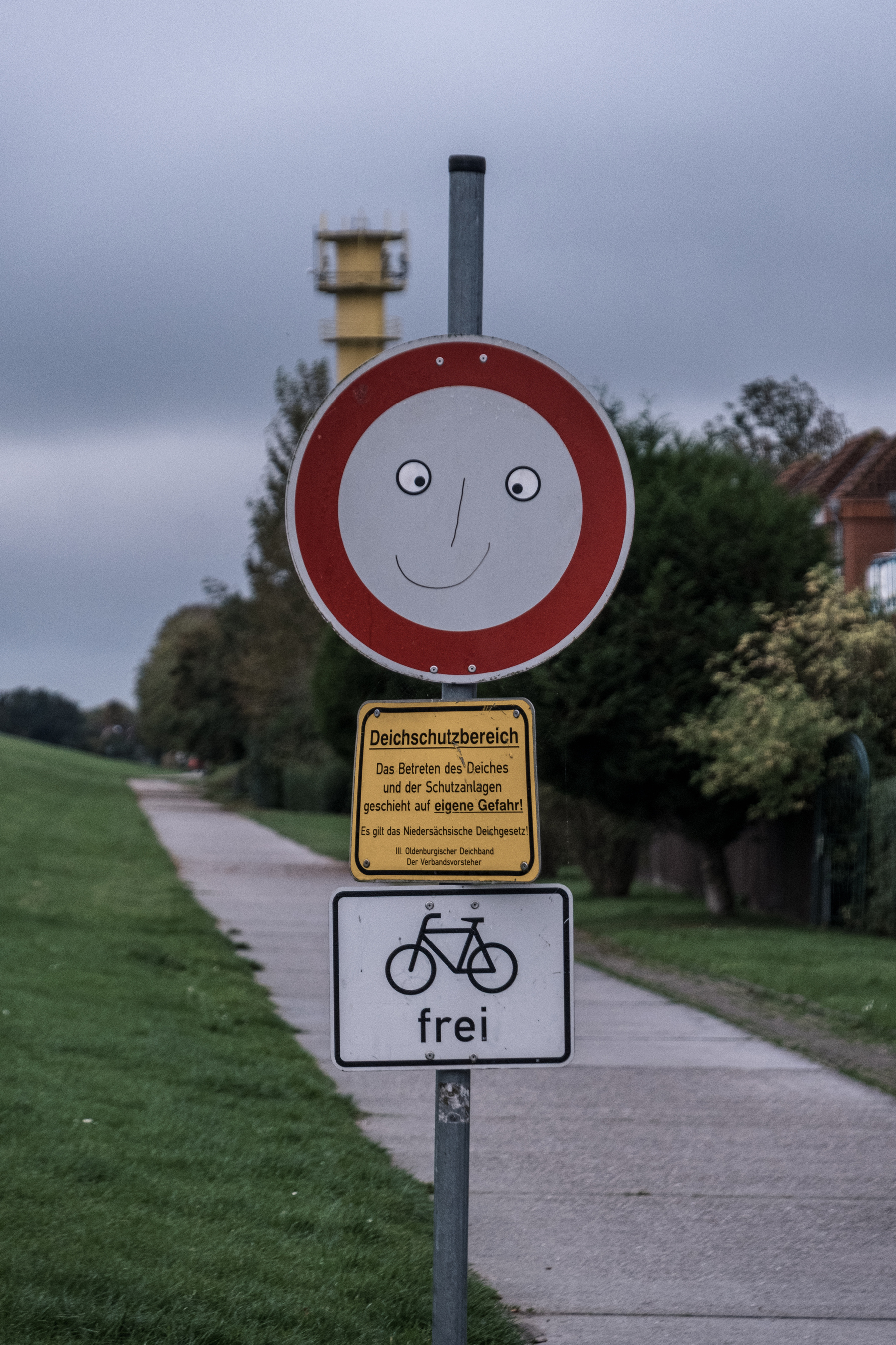 Ein humorvolles Verkehrsschild mit einem Smiley-Gesicht ist über einem Warnschild in deutscher Sprache über das Betreten eines Deichschutzgebiets positioniert, mit einem Fahrradschild mit der Aufschrift "frei". Ein Weg und eine Grasfläche sind im Hintergrund sichtbar.