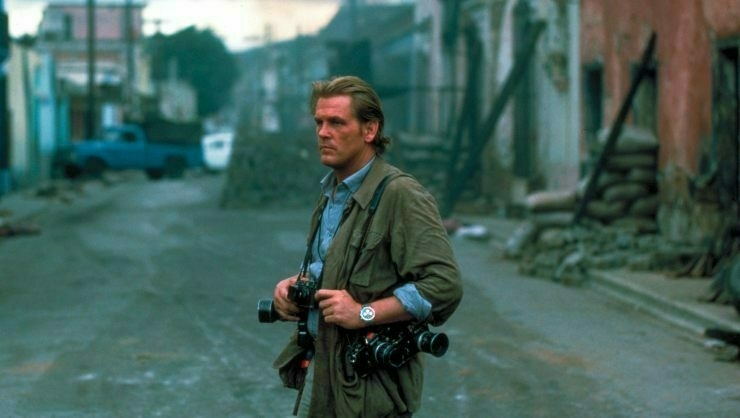 Nick Nolte in "Under Fire" standing on street carrying several cameras