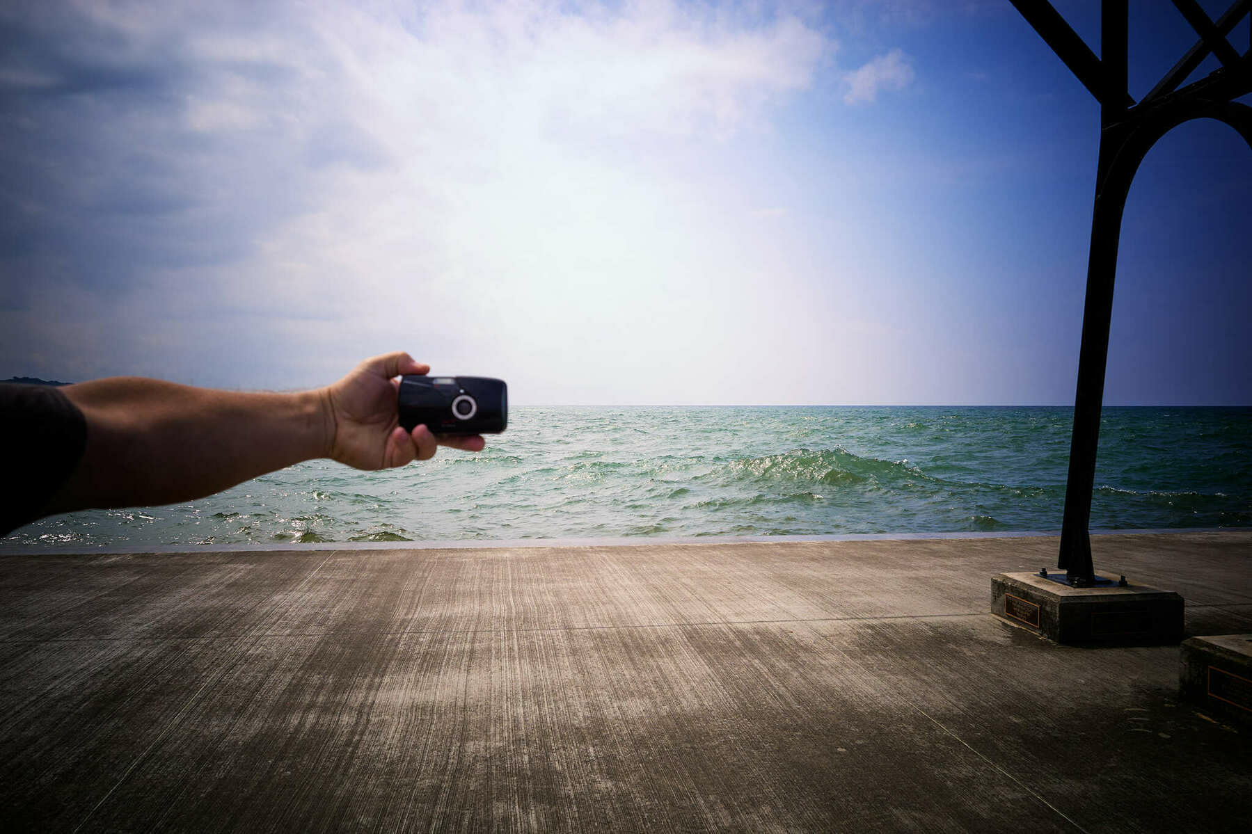 Film camera in outstretched arm pointing at taking camera. Water in background.