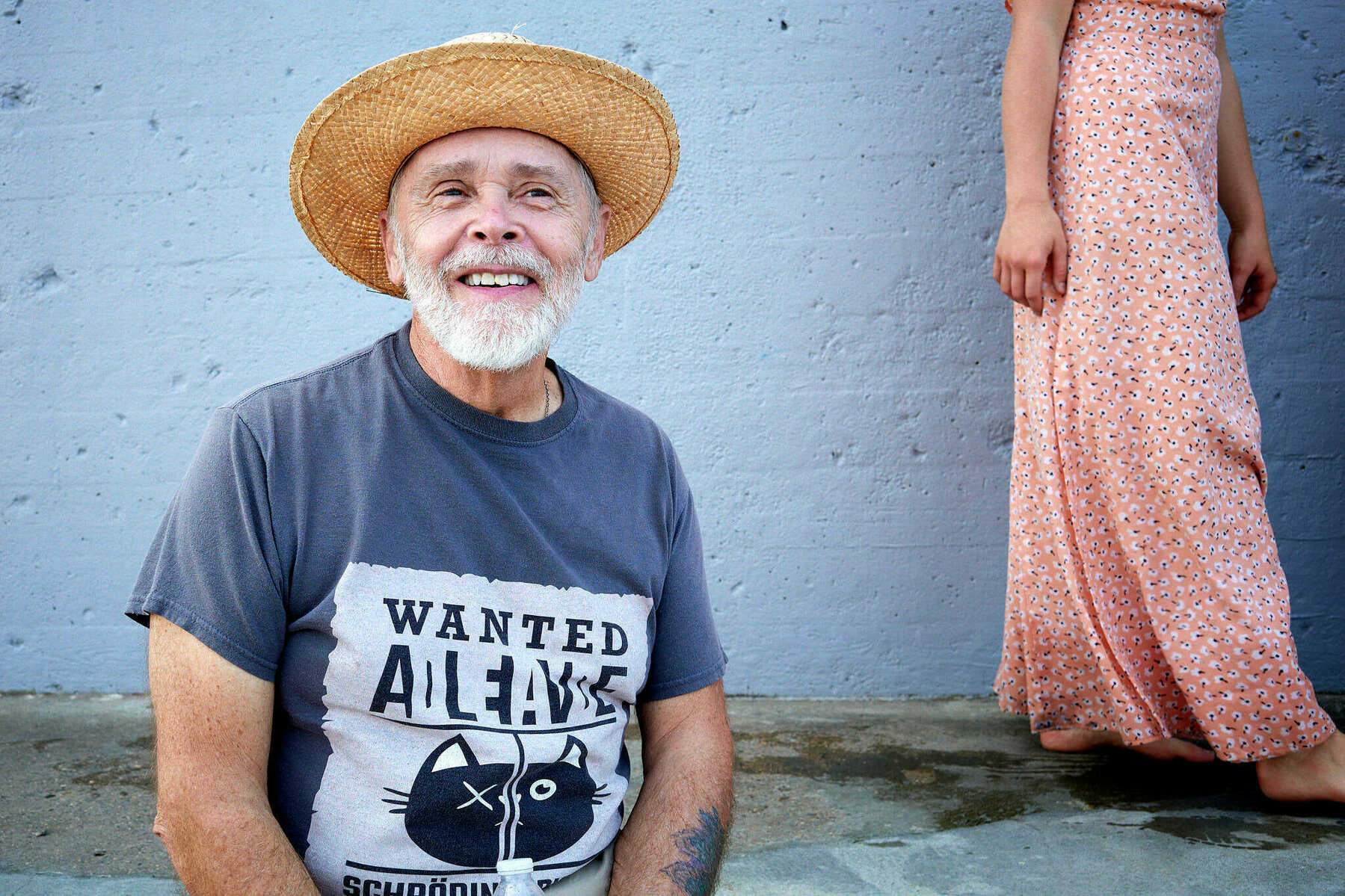 Man in hat sitting in front of a wall with a woman in pink dress walking behind him