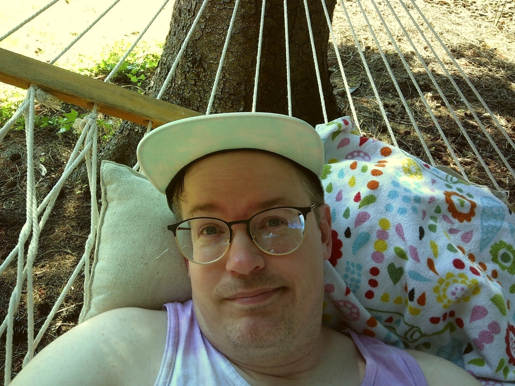 a selfie of a white guy in a tanktop and 5 panel hat who is in a hammock outside in the sunshine