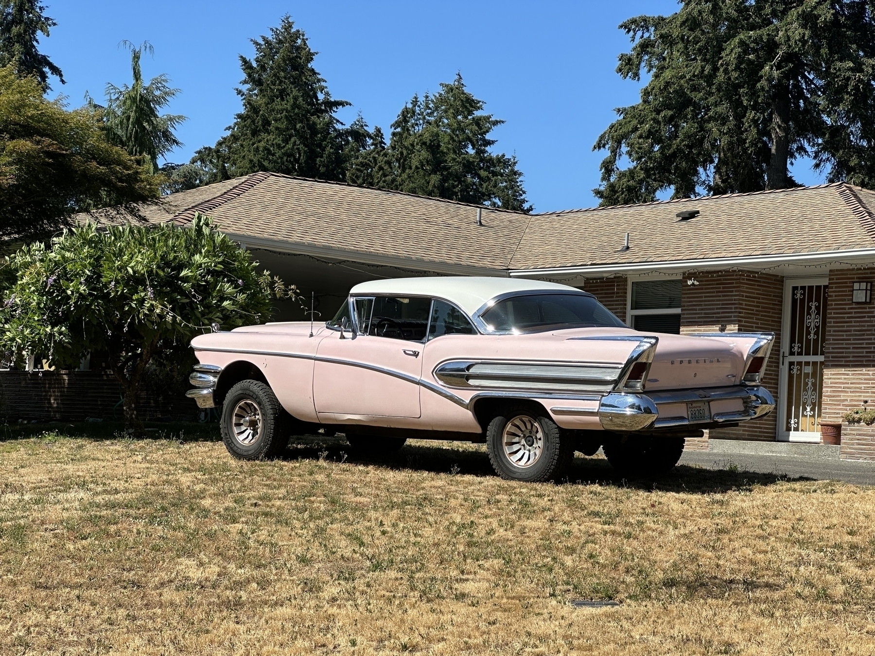 A very old pink car with fins and chrome outside in the sun in a front yard 