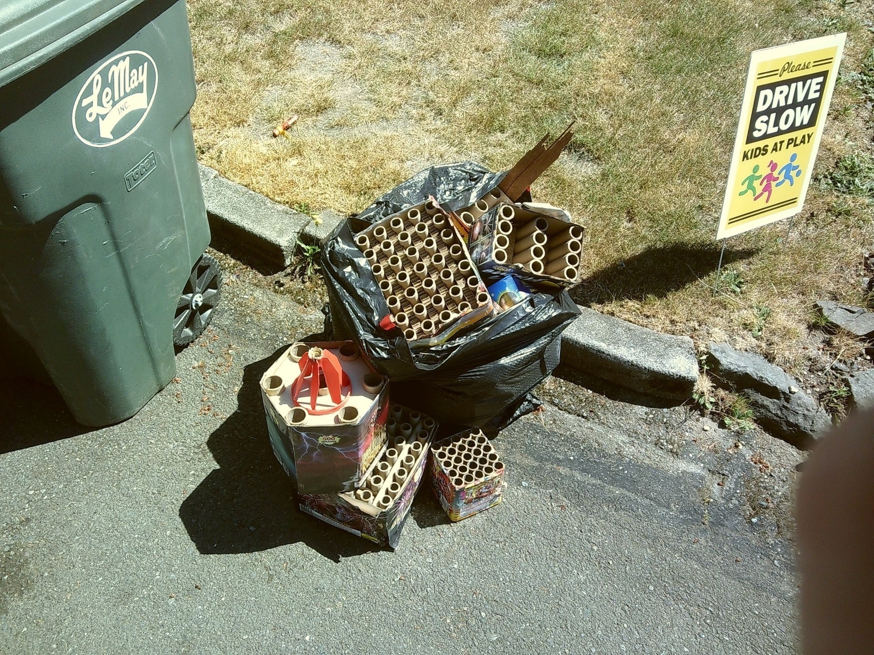 A pile of fireworks debris sits on the road next to a green garbage can and a sign that says Please Drive Slow: Kids at Play