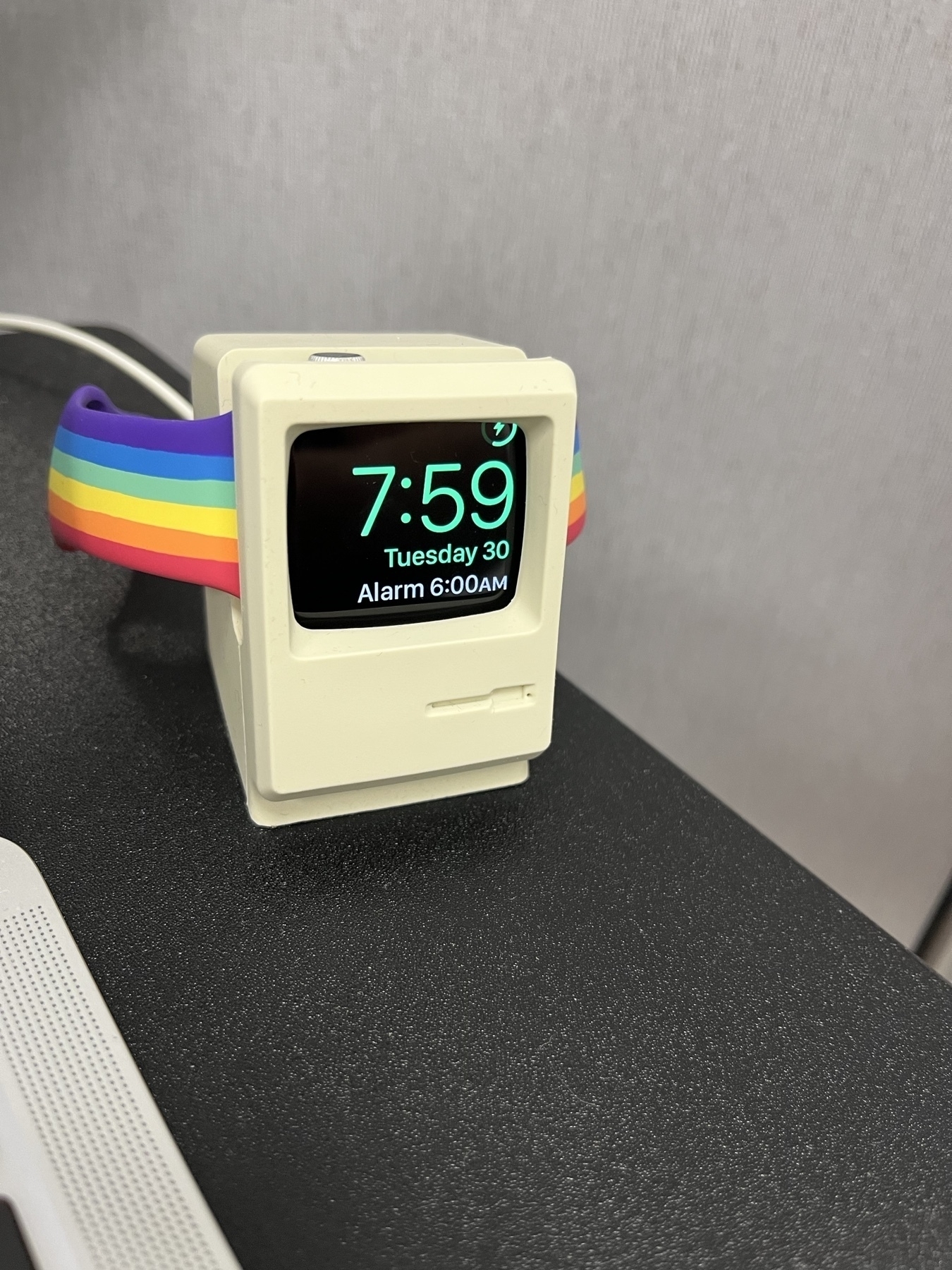 An apple watch with a pride armband is in clock mode in a stand that looks like an original Macintosh.
