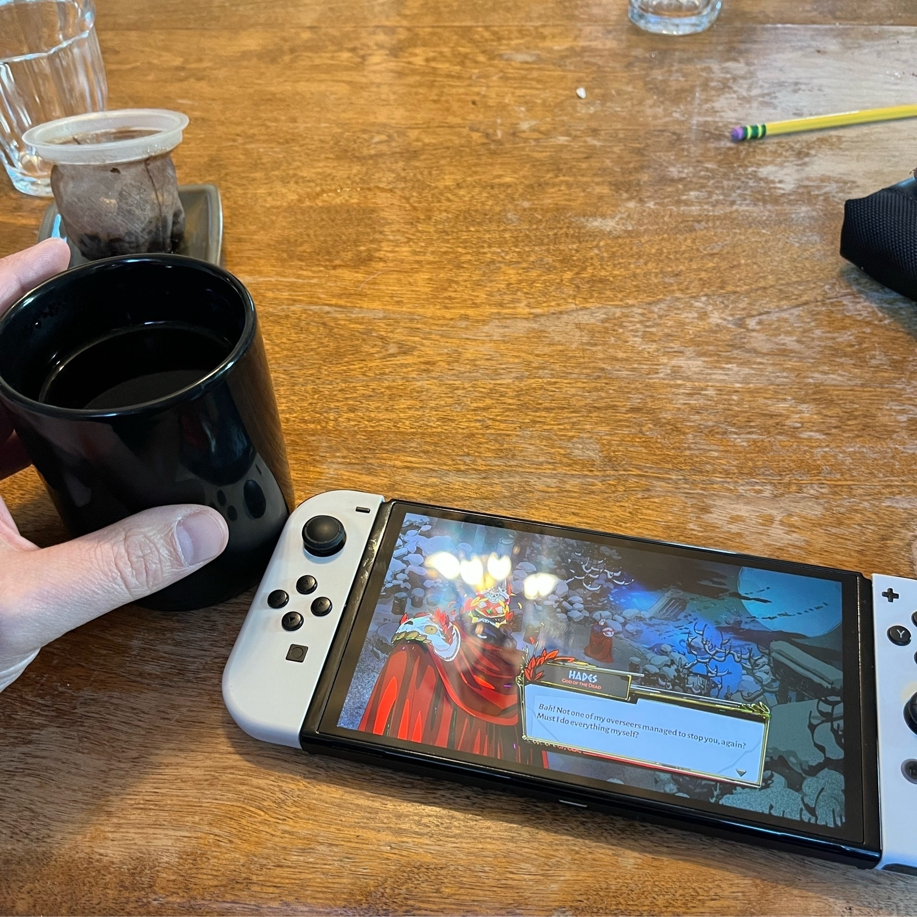 A hand holding a mug of Earl Grey tea next to a white OLED Nintendo Switch playing Hades.