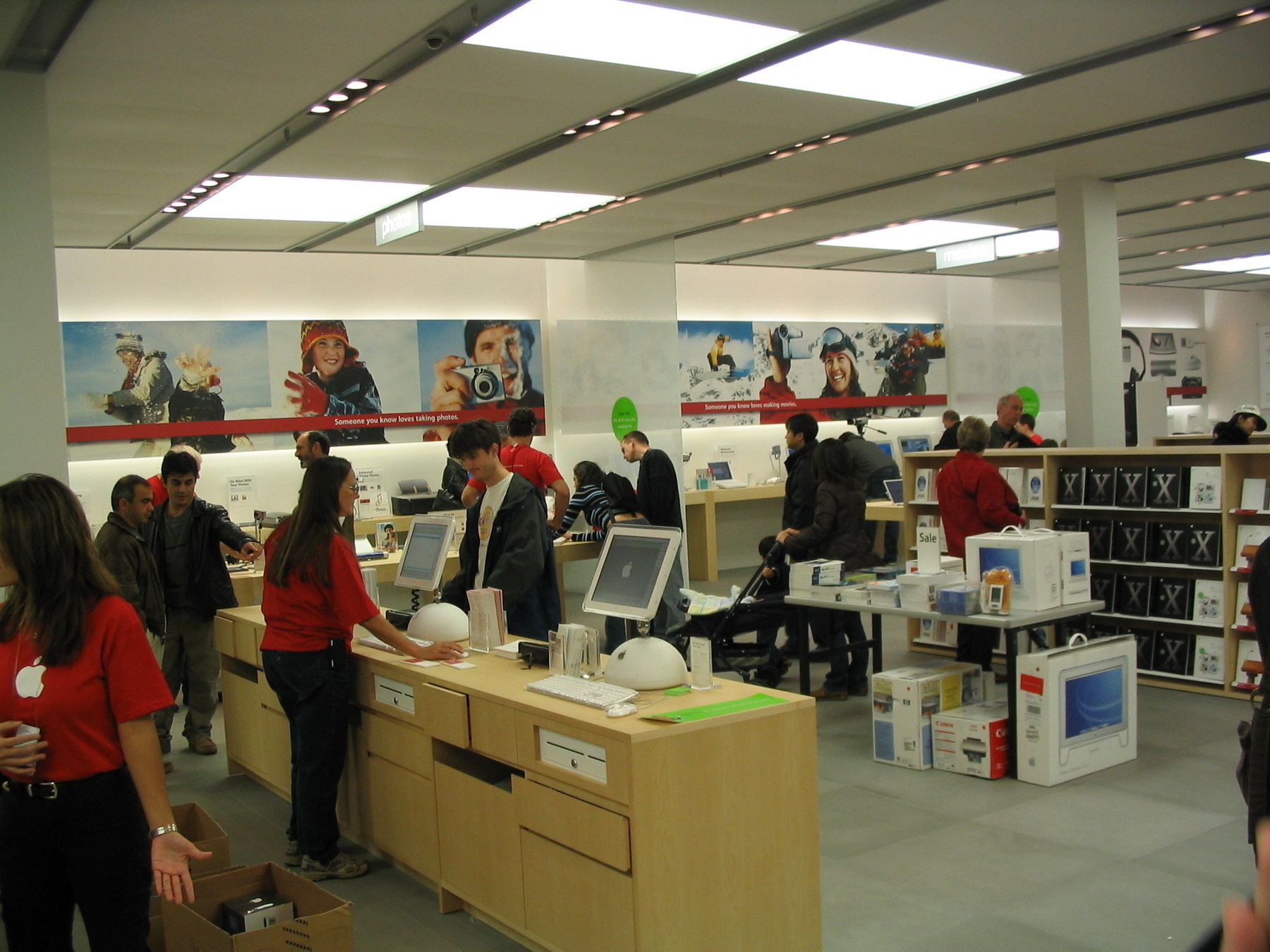 Apple Store workers in red t-shirts with white Apple logos take care of customers at cash registers controlled by iMac G4s.