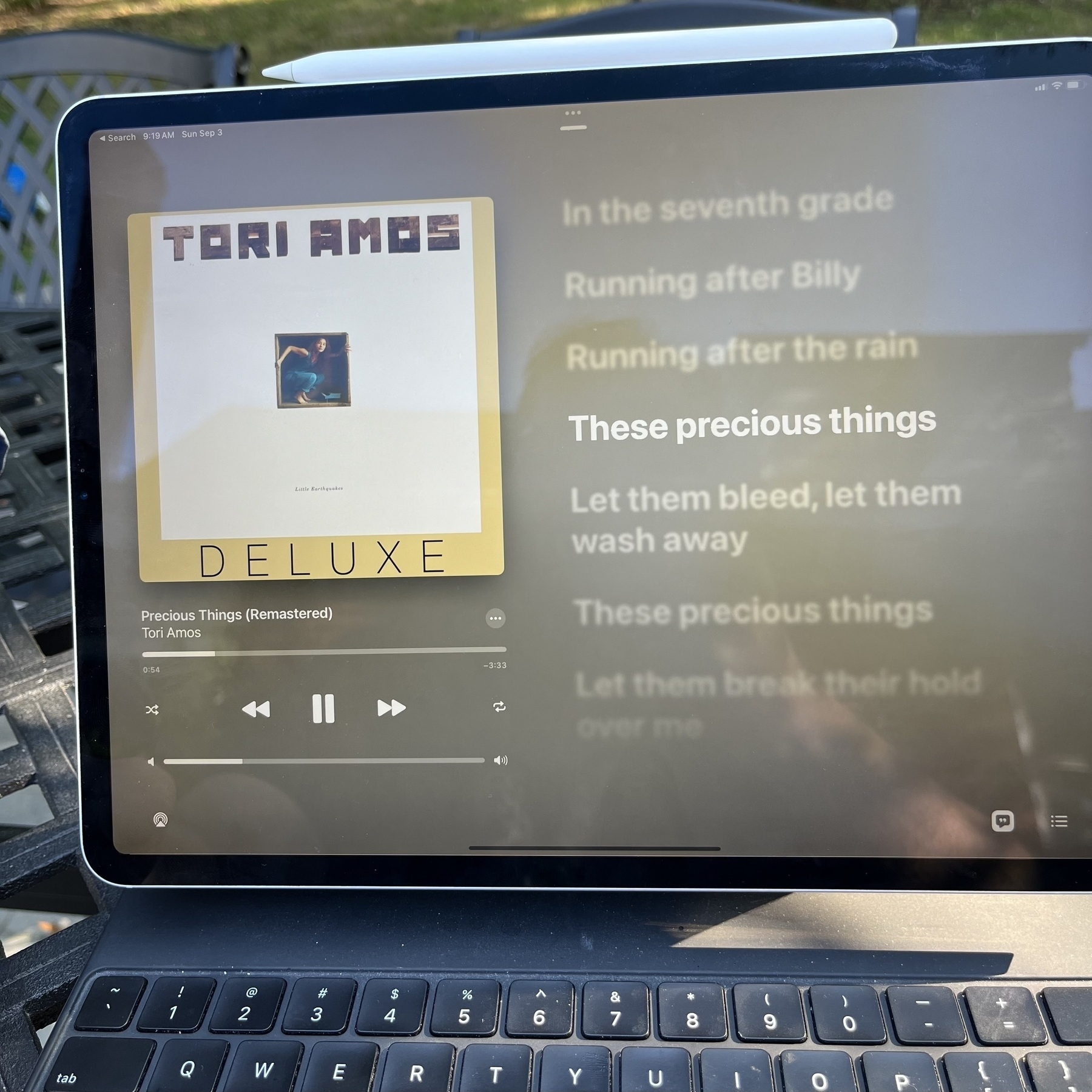 An iPad Pro attached to a Magic Keyboard with an Apple Pencil 2 on top displaying the Music app playing Precious Things from the Tori Amos album Little Earthquakes. The album art is displayed on the left, with Music.app’s controls and the lyrics are displayed on the right with the line “These precious things” highlighted.
