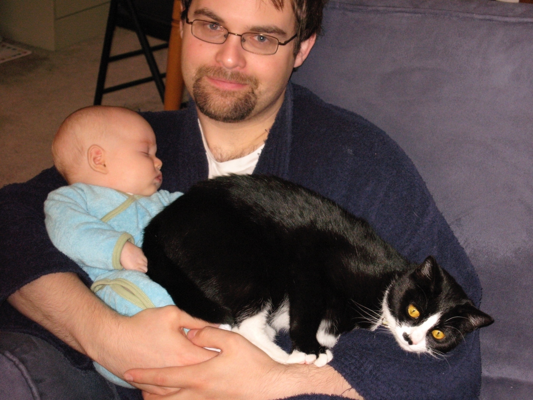 Sam holds an infant while a cat sits on the infant.