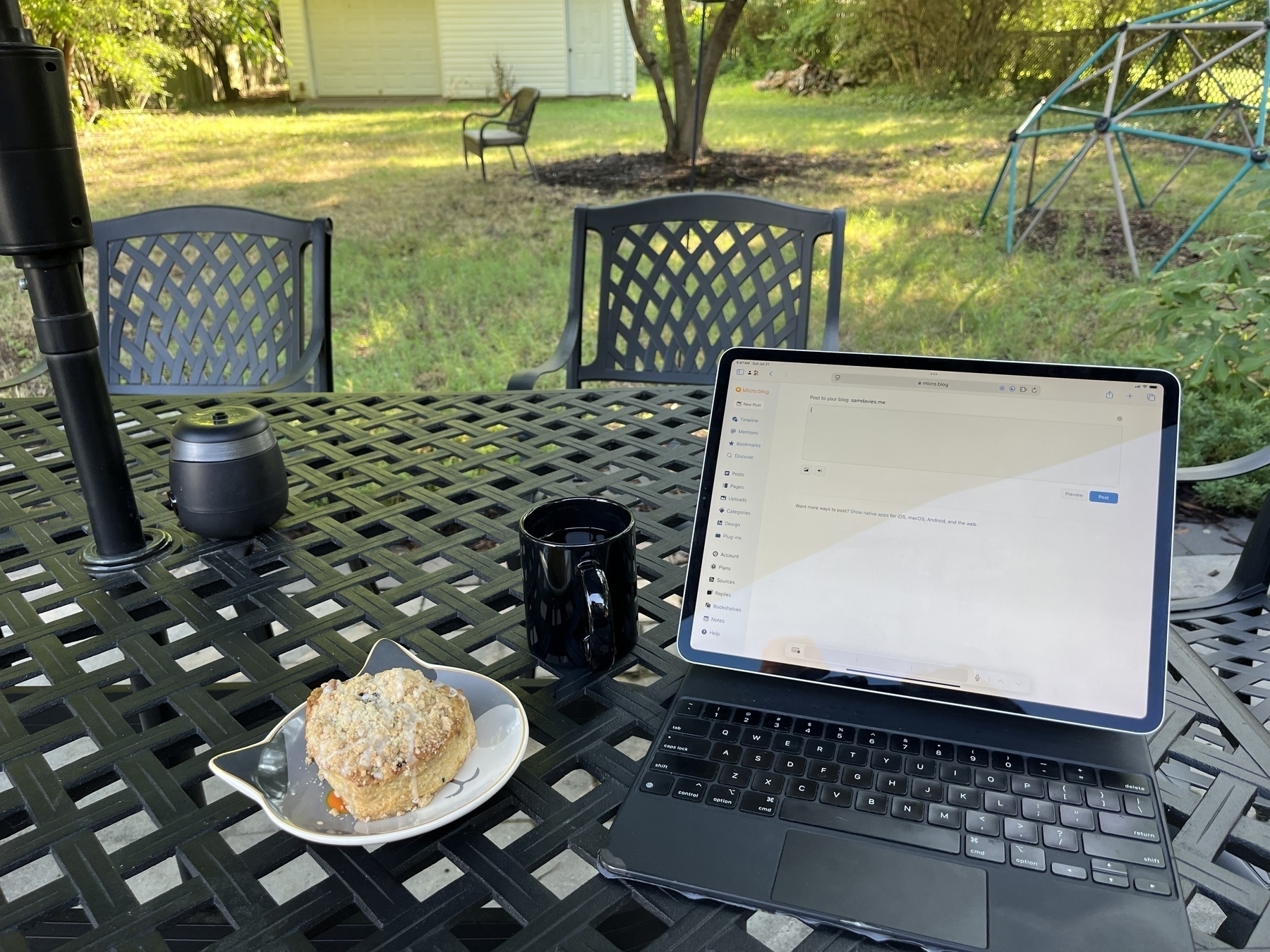 A metal patio table with a scone on a kitty plate. A black mug of tea. An iPad in a keyboard case open to micro.blog.