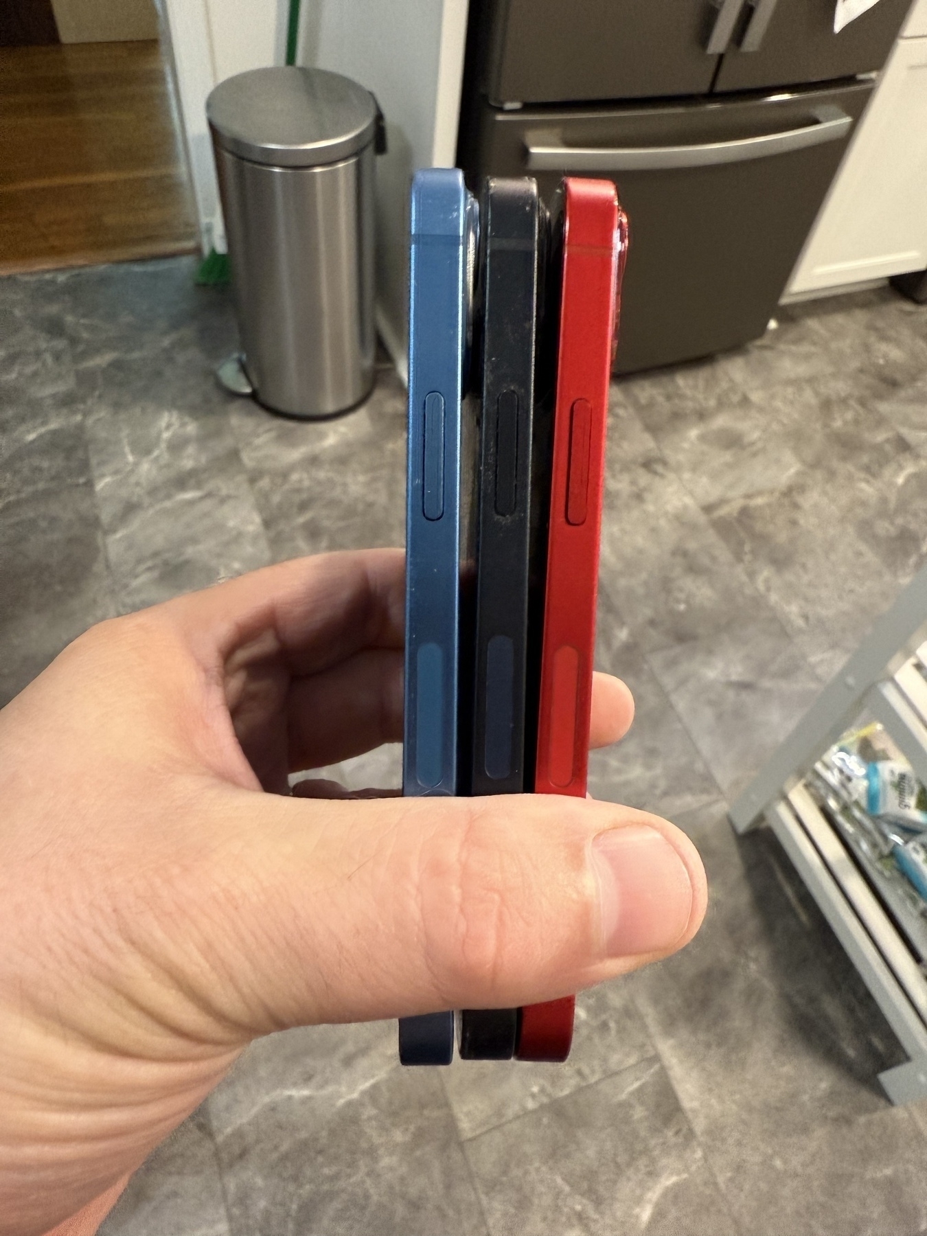 Three iPhone 13 minis in blue, black, and red are held in a hand against a kitchen backdrop.