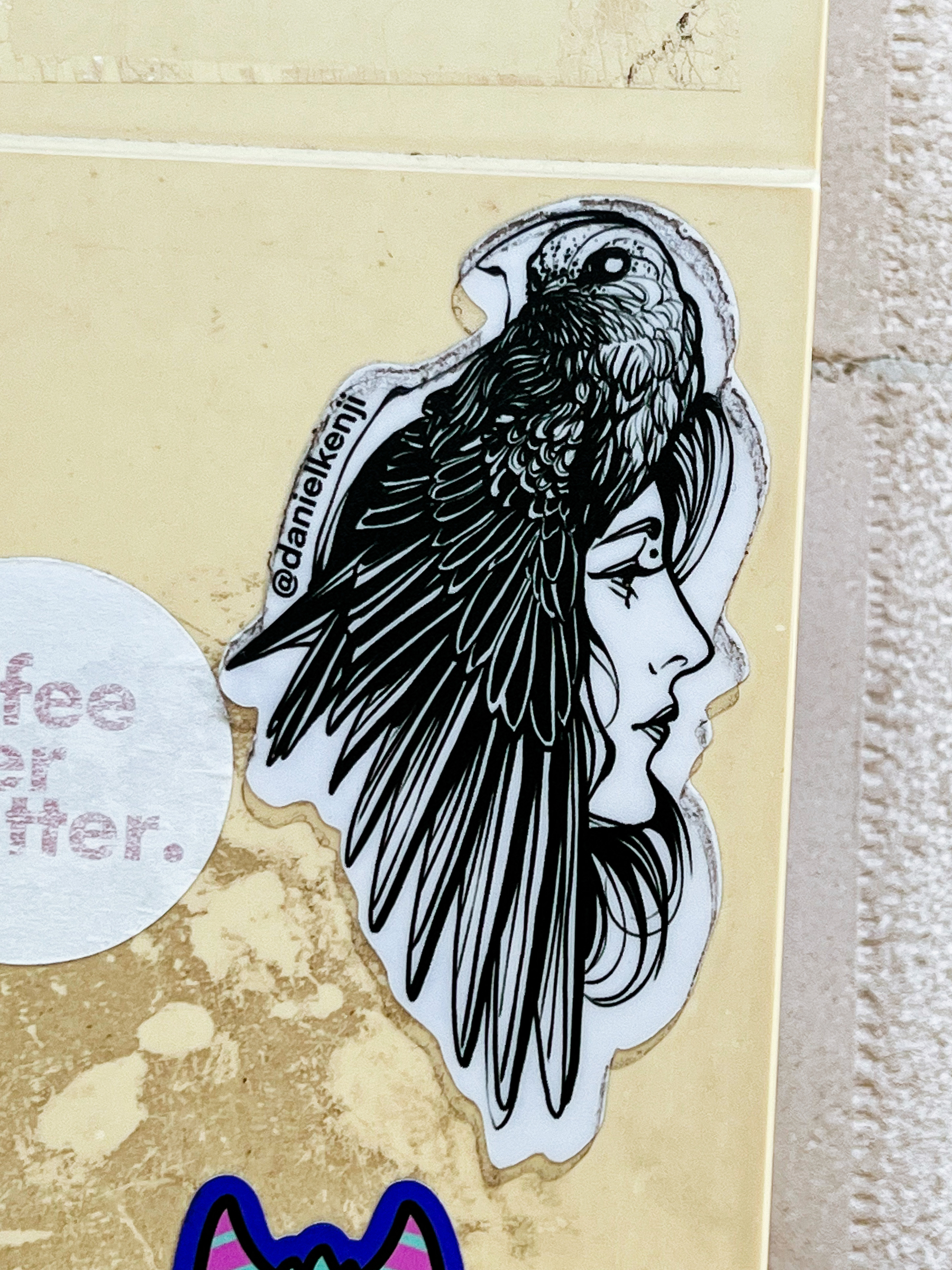 Sticker of a woman’s face. A bird sits on top of her head, his wings make up her hair. 