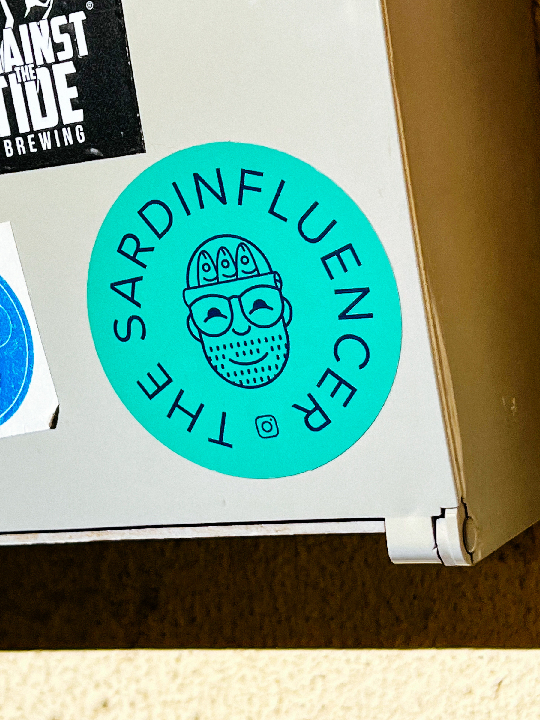 Sticker with the words “The Sardinfluencer”, and a drawing of a bearded man with glasses, and a beanie with three sardines sticking out. 