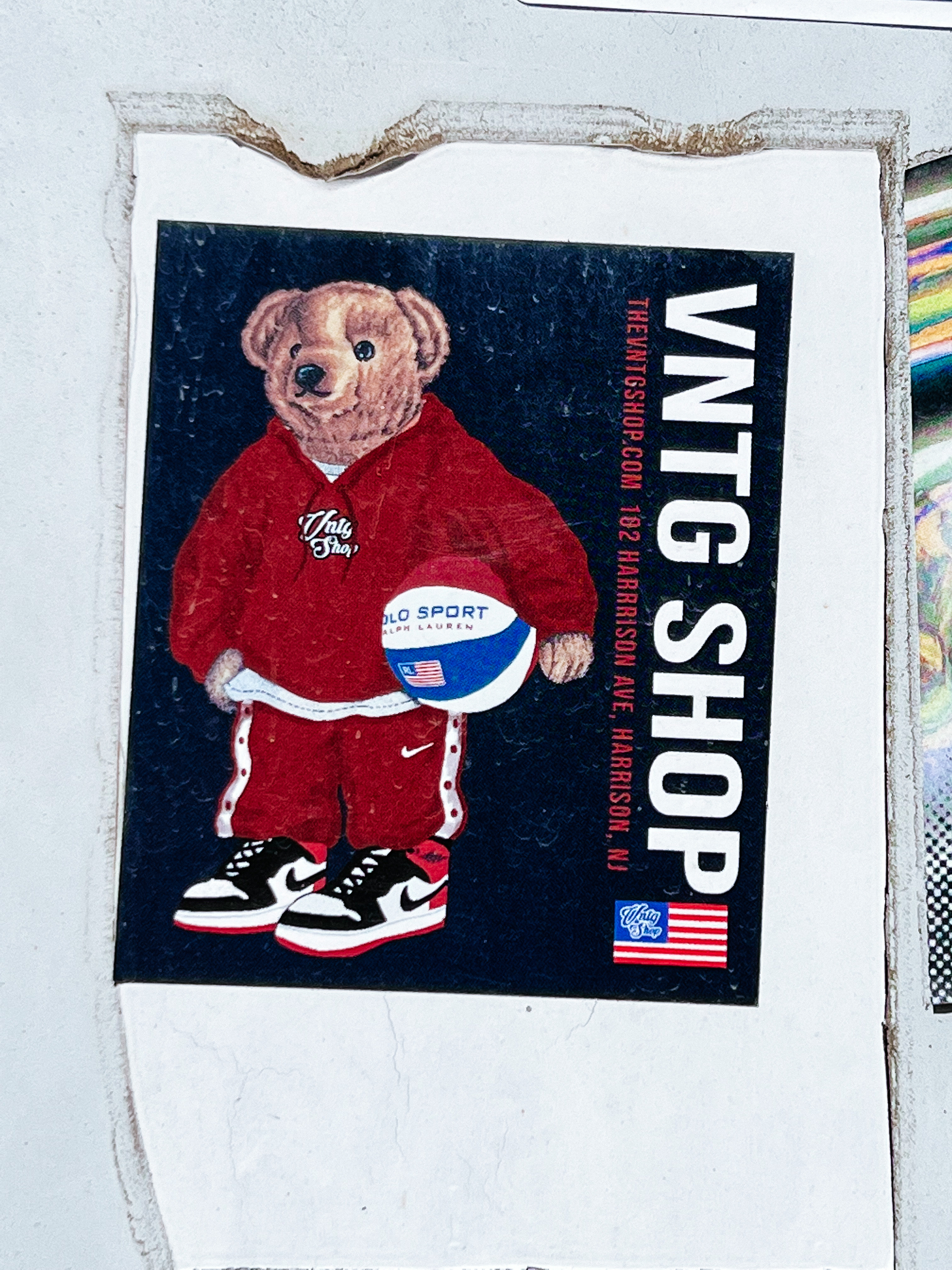 Sticker of a bear in a tracksuit, holding a basket ball. “VNTG SHOP” written on the sticker, with a USA flag. 
