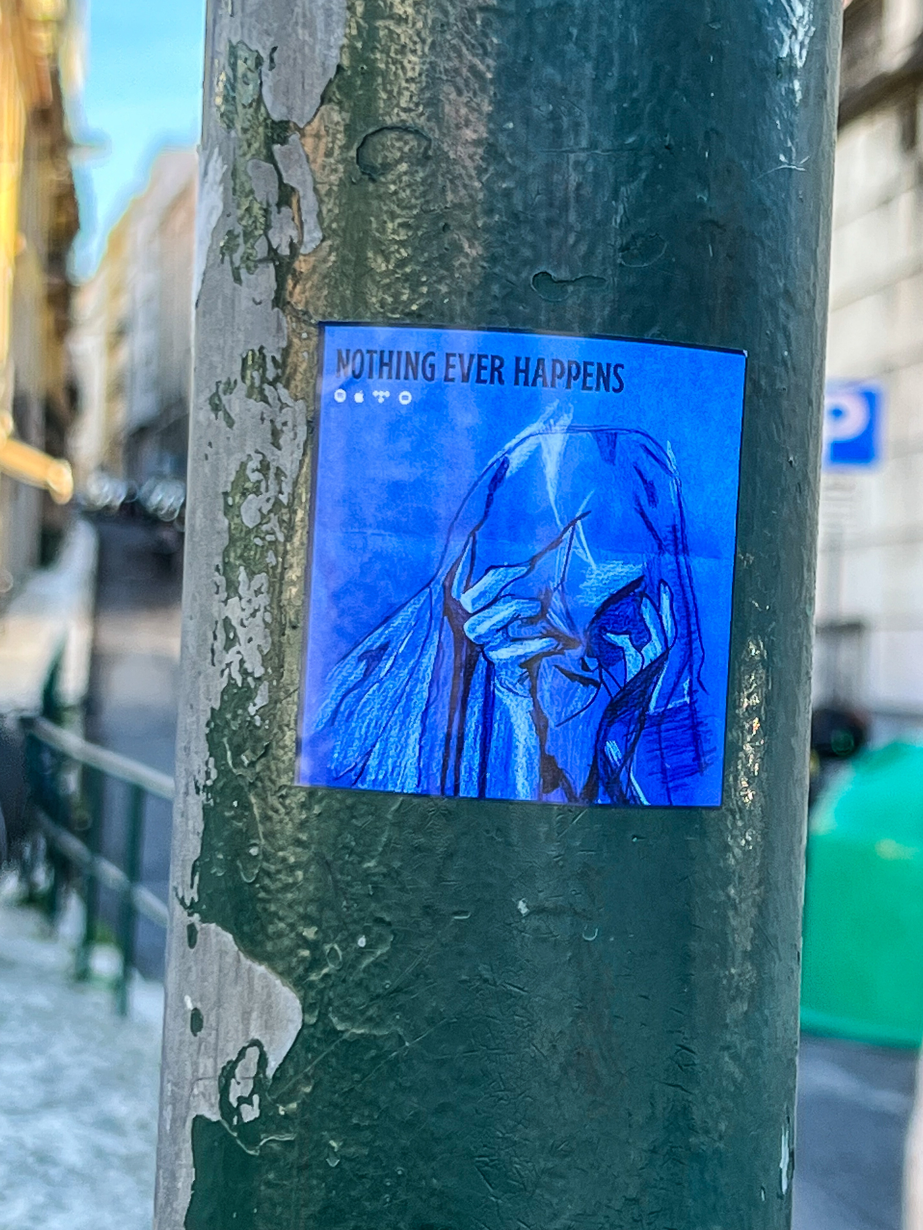 Blue sticker, drawing of hands on a face, with the words “nothing ever happens” written above it. 