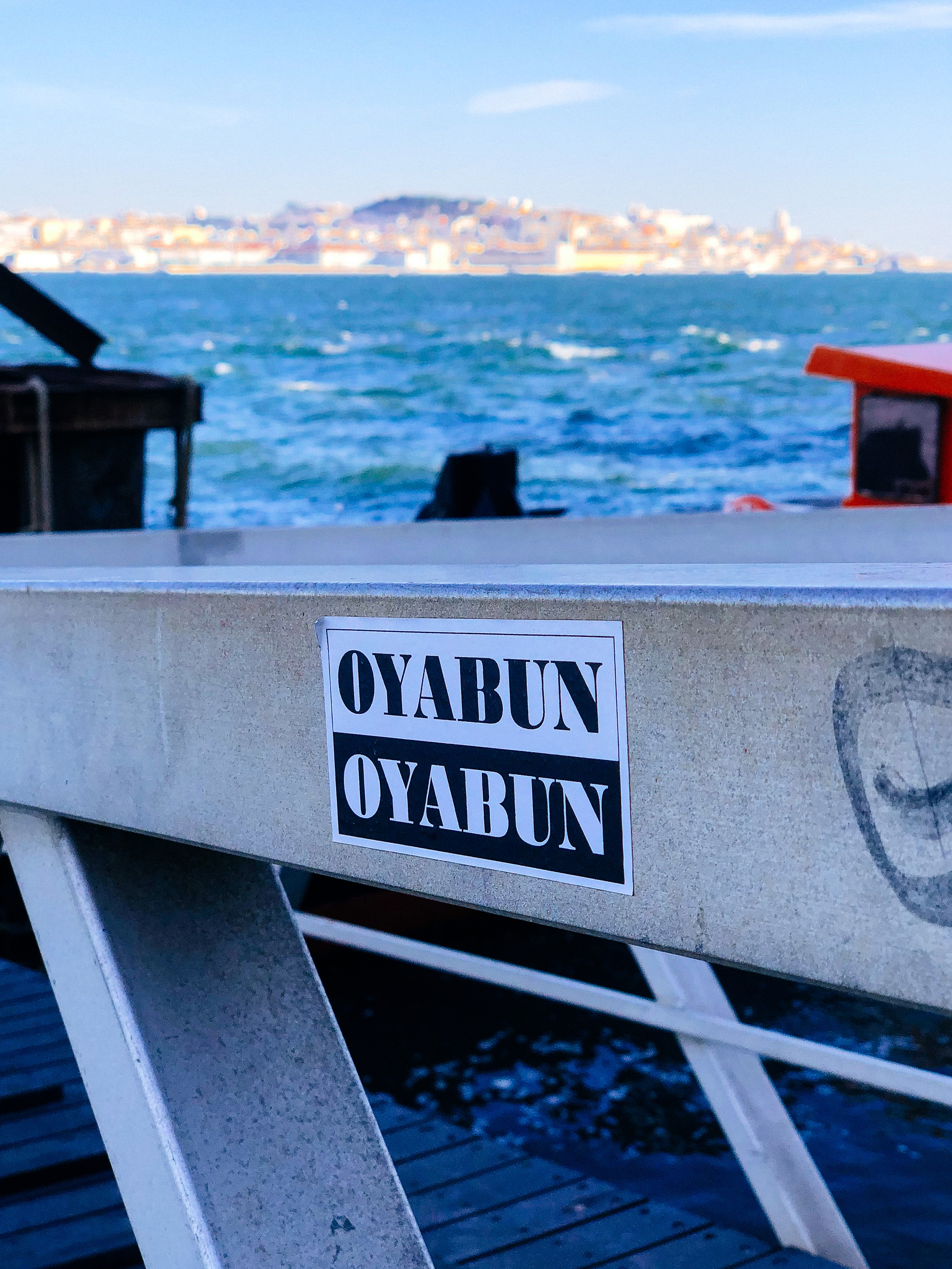 Sticker of the word “OYABUN” written twice. Once black on white, and another white on black. One on top of the other. You can see a river on the background, with a city in the distance, on the other bank. 