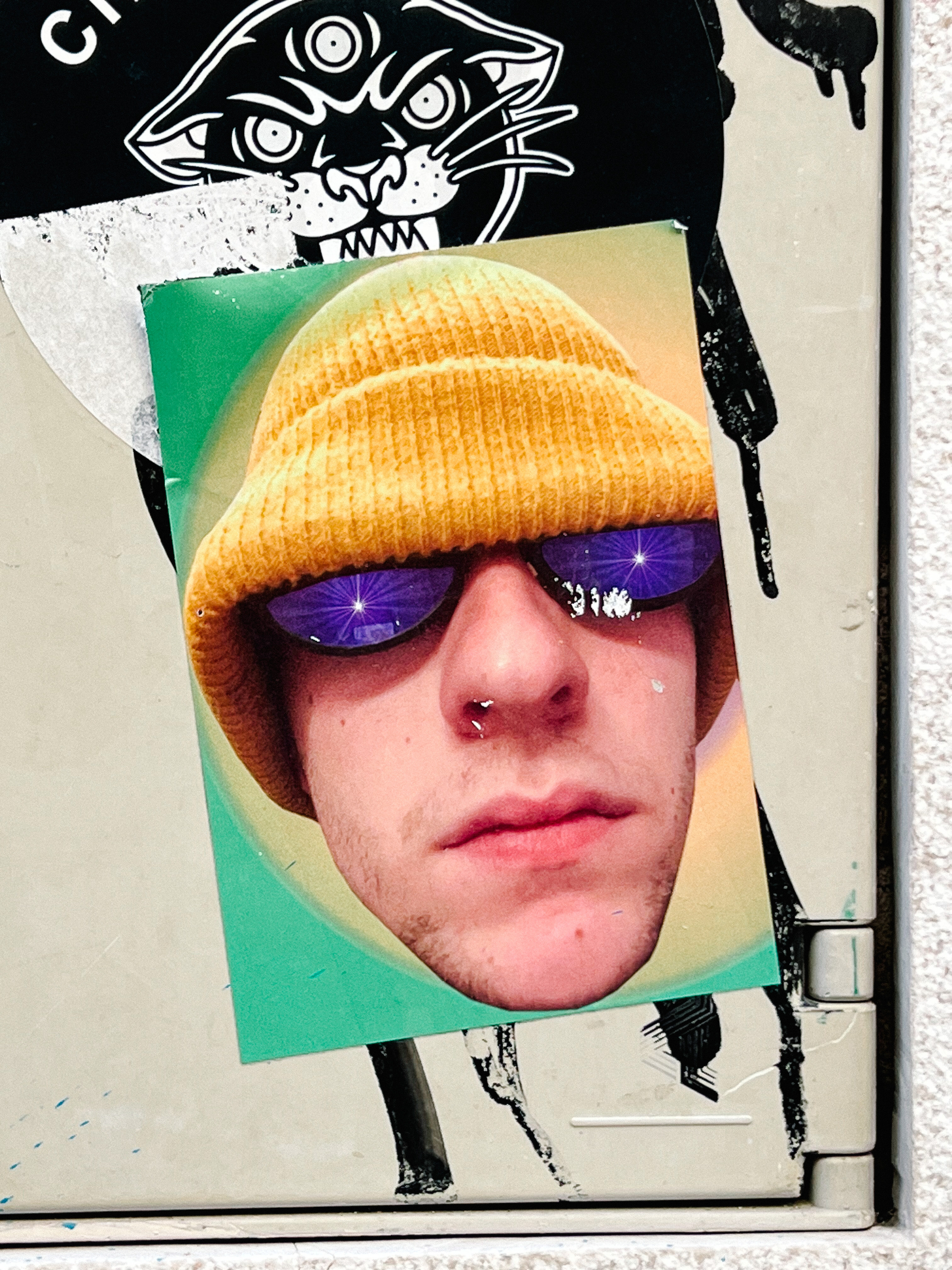 Sticker with a photo of a man’s face. Wearing shades, and a yellow beanie. 
