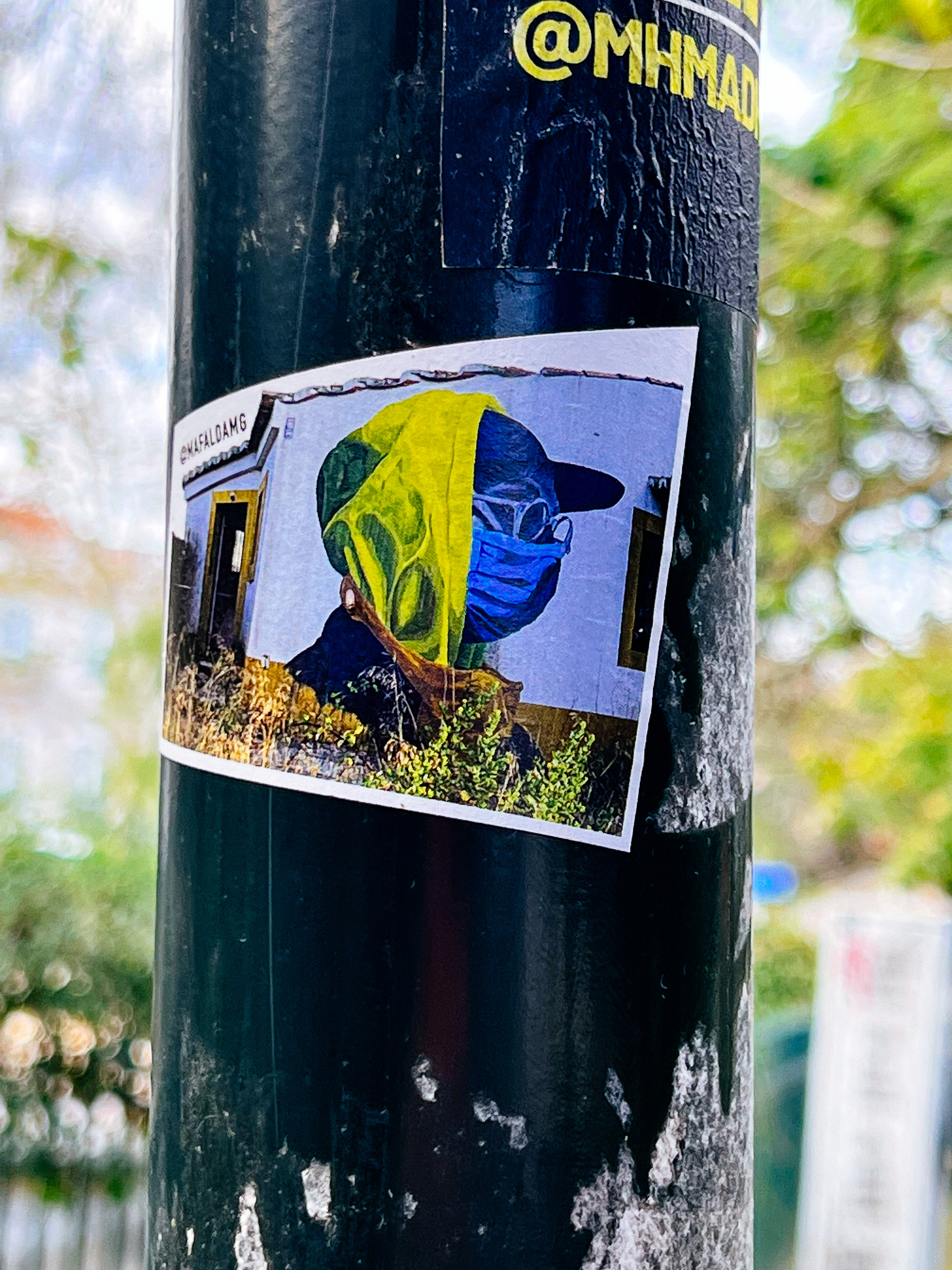 Sticker with the photo of a wall with a street art piece. There’s a man wearing a yellow scarf, and a blue mask, Ukrainian colors. 