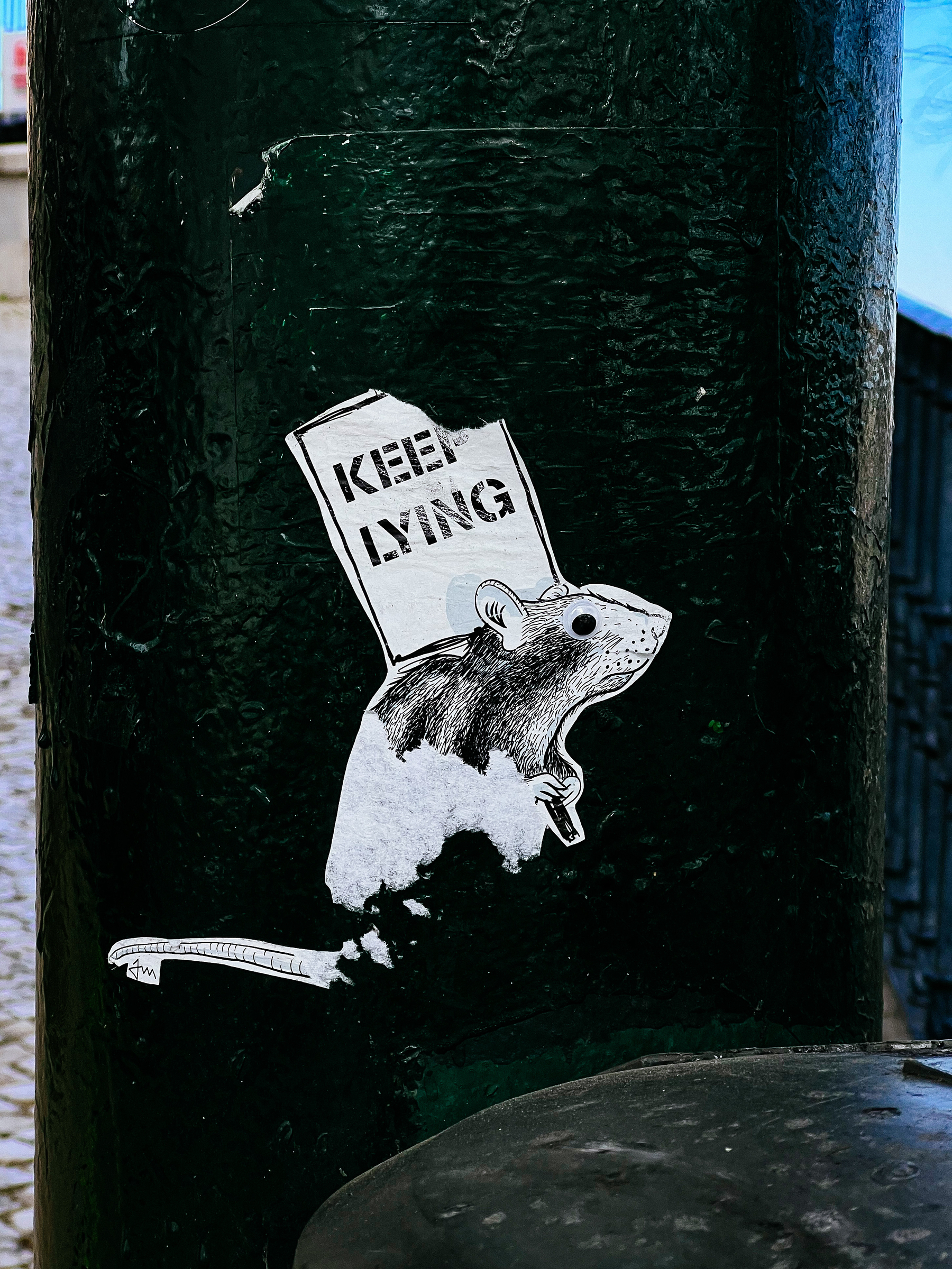 Sticker of a mouse holding a sign that says “Keep Lying”. 