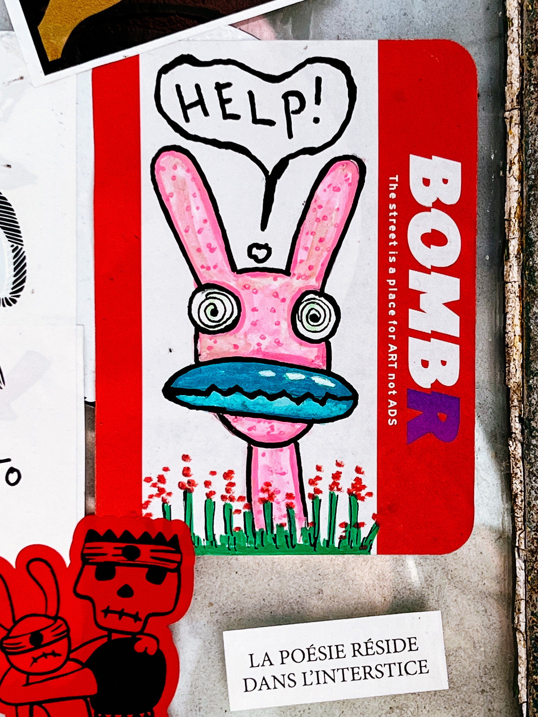 Sticker combo. A mess. There’s a cartoon creature, sorta like a rabbit, a pink one, with crazy hypnotized eyes, a very large blue mouth, and a cry for “HELP!”. That sticker has “BOMBRThe street is a place for ART not ADS.” written on it. Below you’ll find a simple black text on white background, with “LA POÉSIE RÉSIDE DANS L’INTERSTICE”. Still place for an old friend, the skull faced girl with the eye patch wearing bunny.