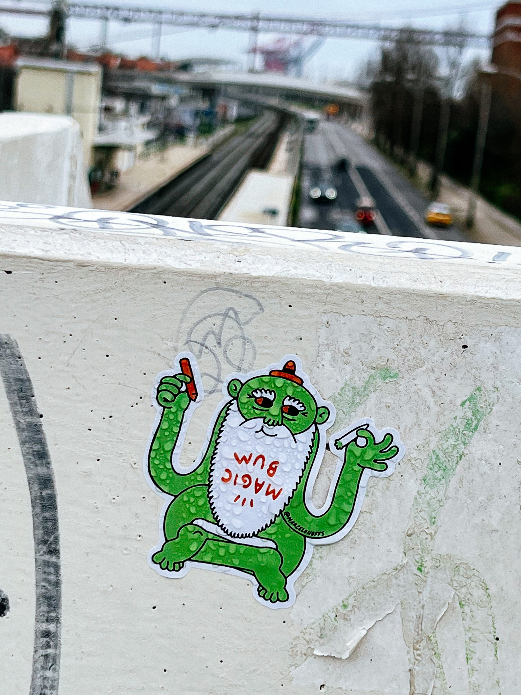 Sticker with a green yoga master, holding a pencil in one hand, and a joint in the other. He has a tiny orange hat, and a big white beard with the words “magic bum” written upside down. 