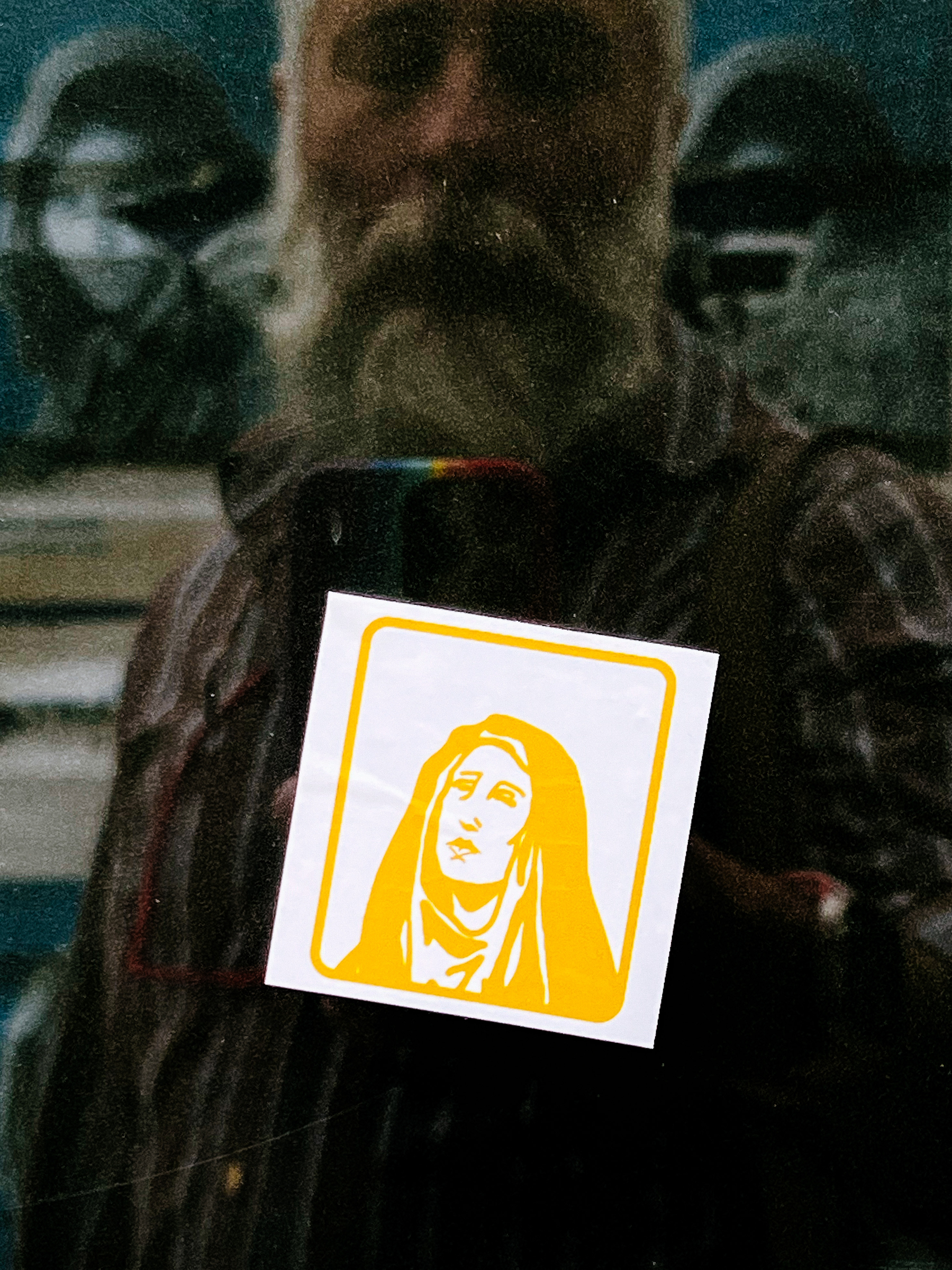 A sticker with an illustration of Mary, mother of jesus. Reflected we can see the photographer. And two of Steve McCurry’s Afghan girl portraits. 