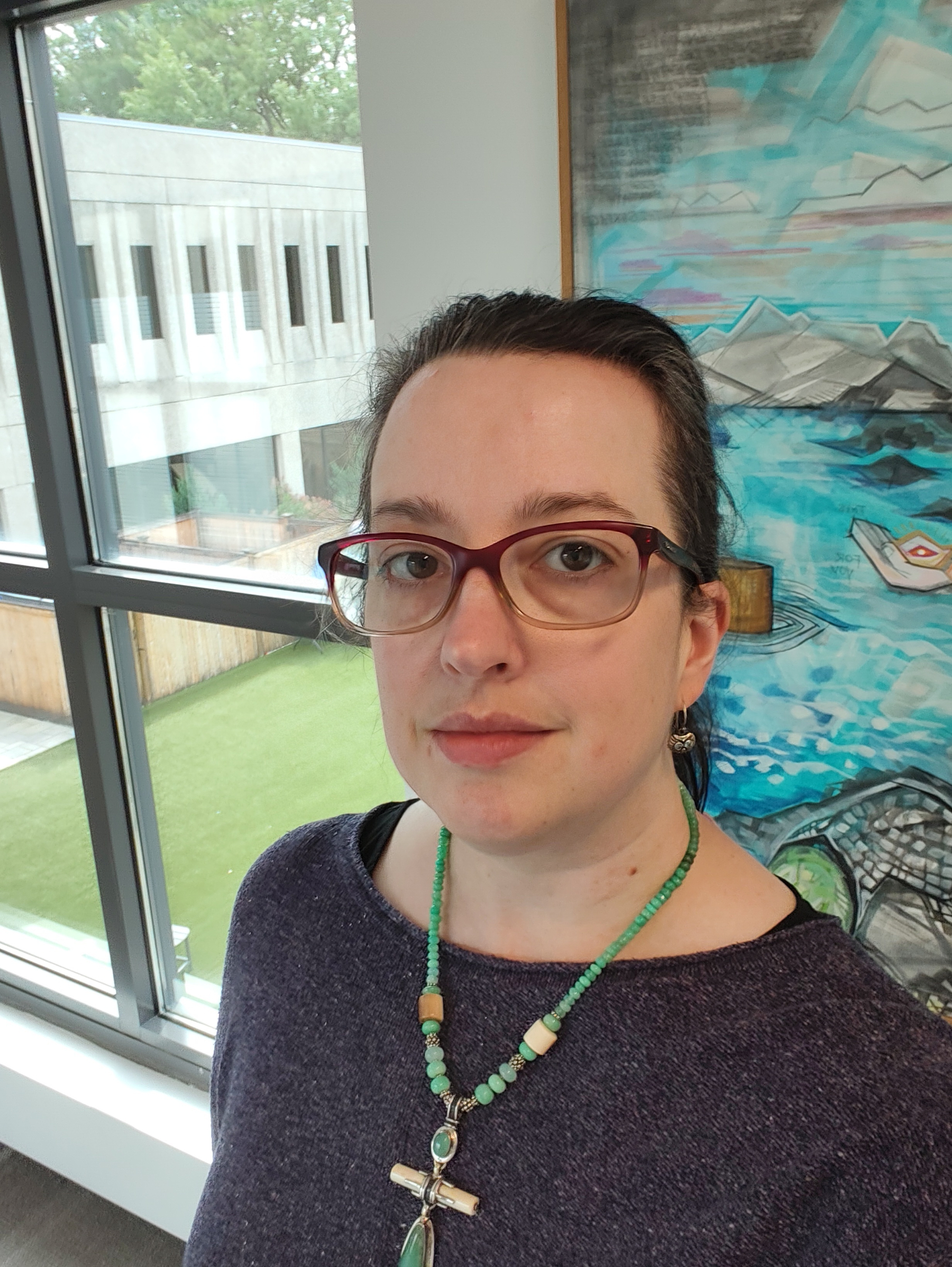 A woman wearing glasses, a purple sweater, and a turquoise necklace is indoors near a large window with a grassy outdoor area and a colorful ocean-themed painting in the background.