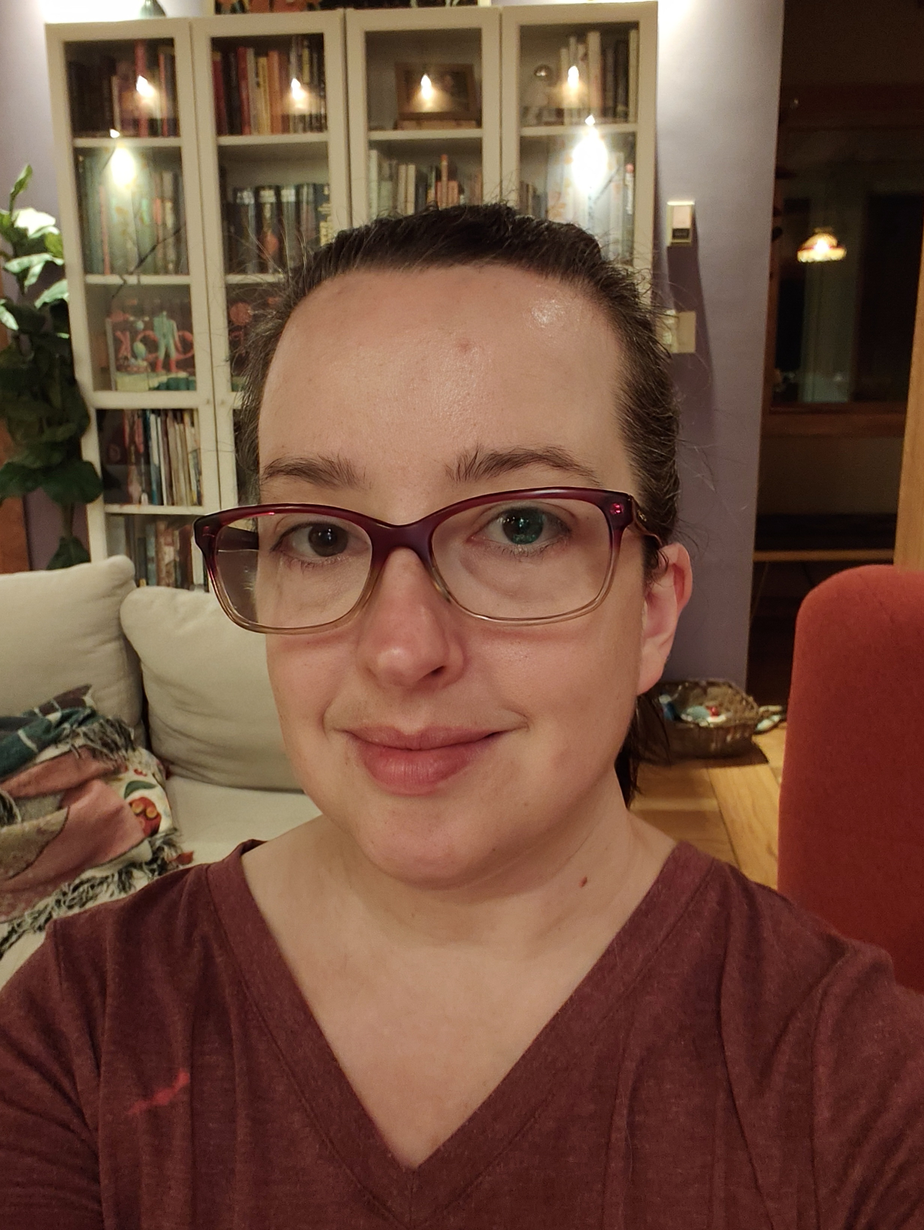 A person with glasses and a maroon shirt is smiling inside a cozy room with bookshelves and a couch in the background.