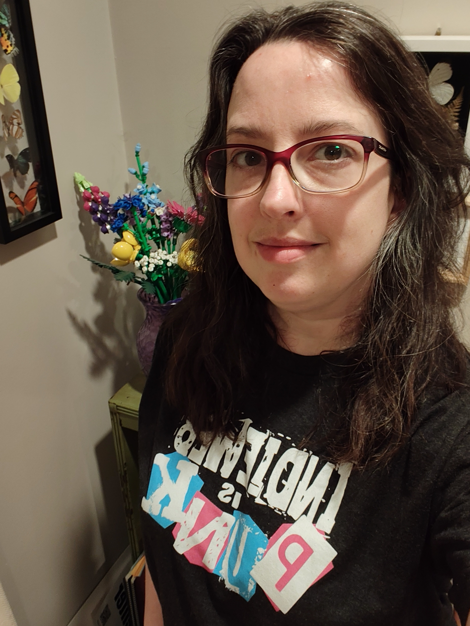 A person with long dark hair and red-framed glasses is smiling slightly while wearing a black t-shirt with colorful text, standing indoors near a vase of vibrant artificial flowers and framed butterfly decorations on the wall.