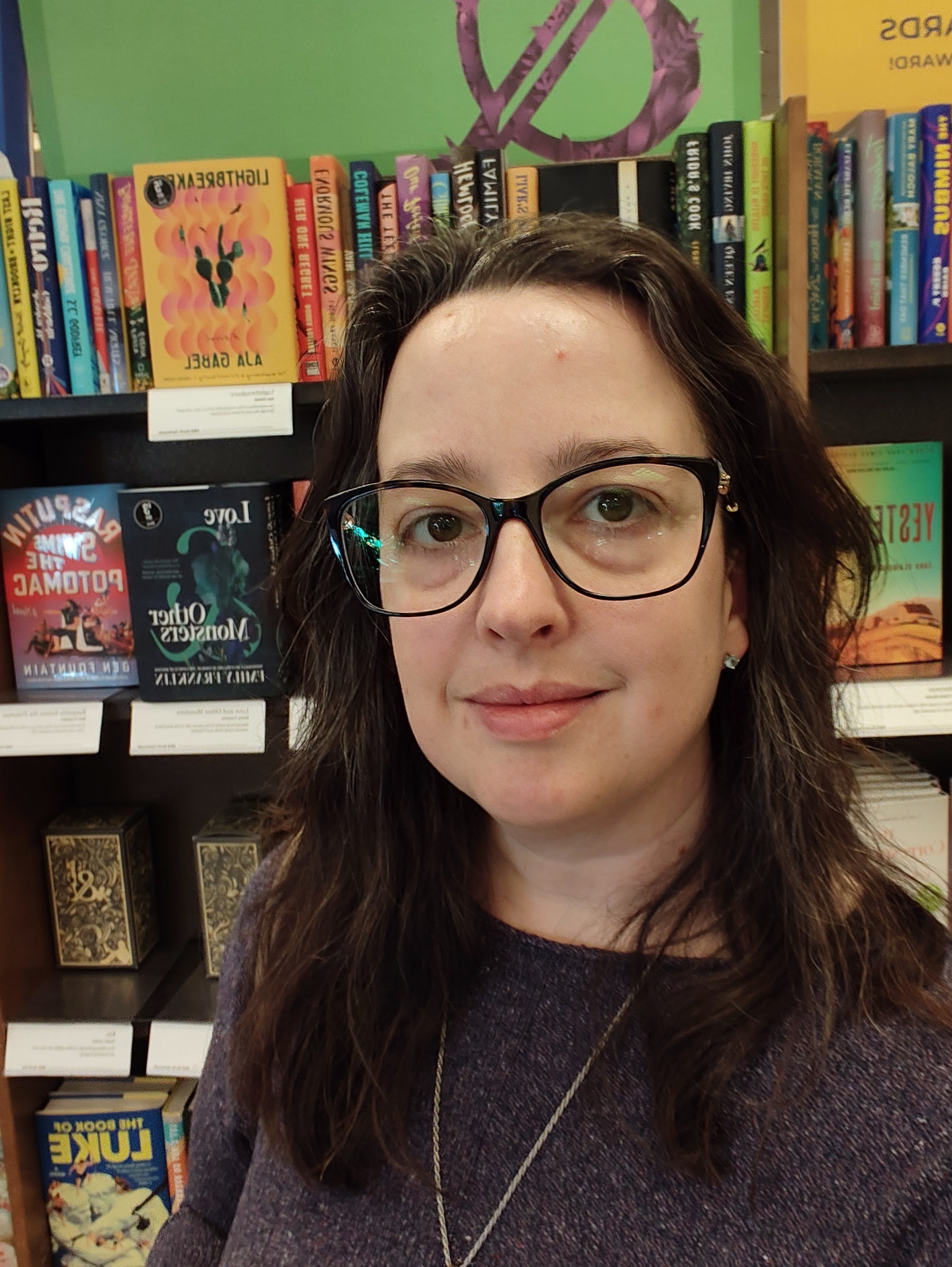 A woman with glasses and long brown hair is smiling slightly while standing in front of a bookshelf filled with colorful books.