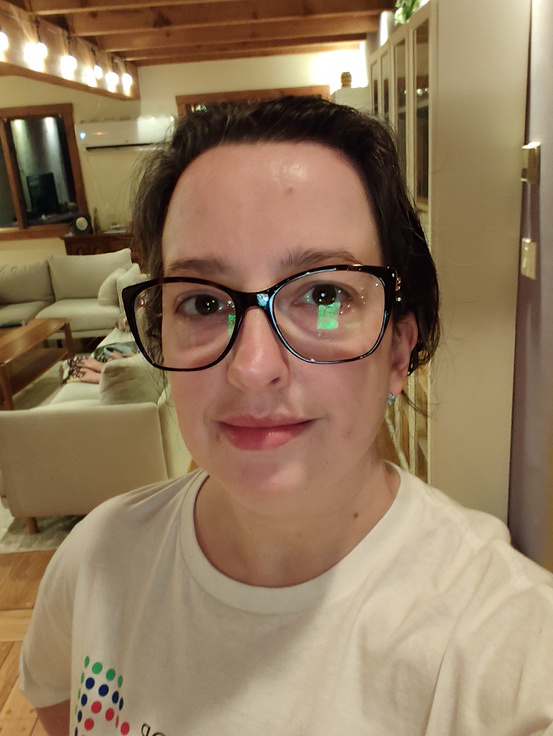 A person wearing large black glasses and a white t-shirt is taking a selfie in a warmly lit room with wooden beams and cozy furniture.