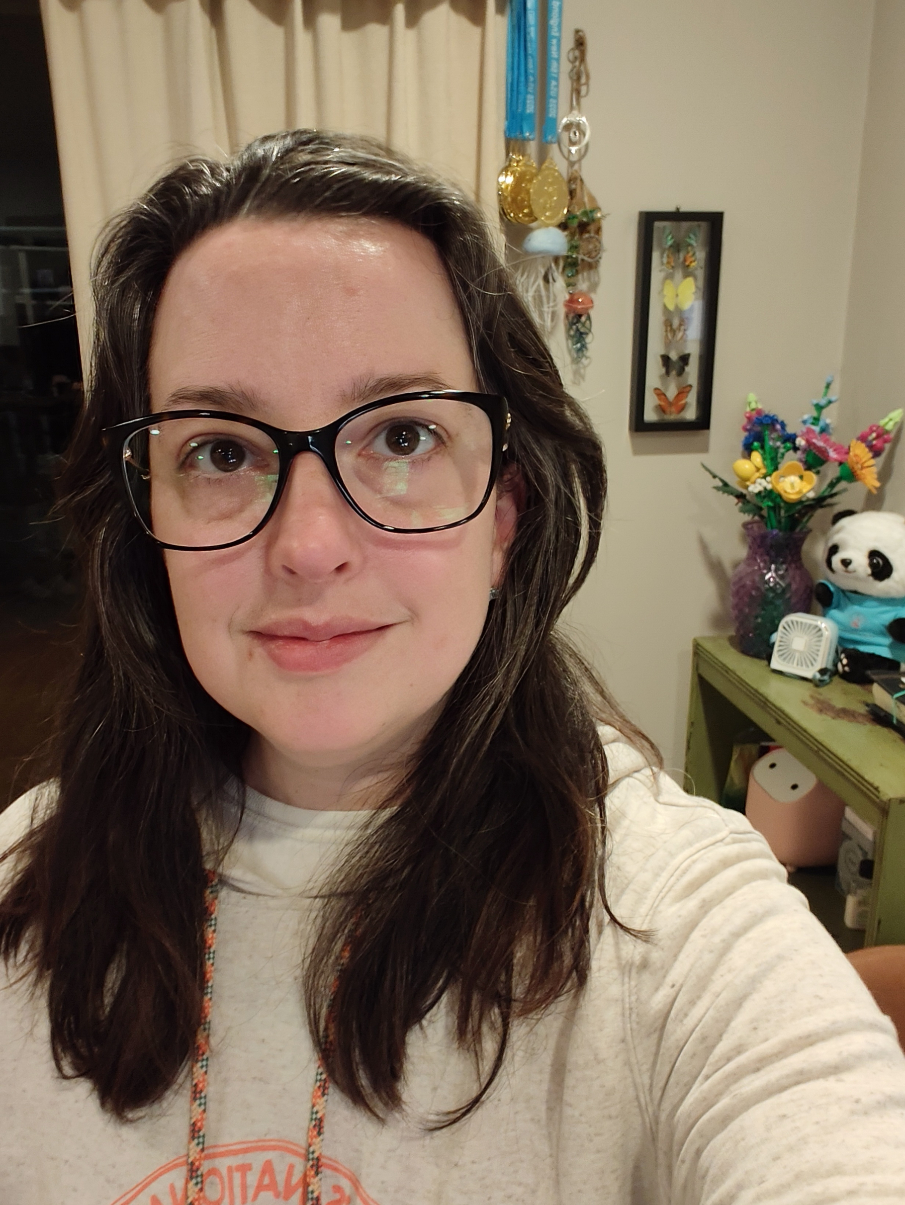 A woman with glasses and long dark hair is smiling softly in a cozy room decorated with colorful flowers, medals, and a panda figurine.