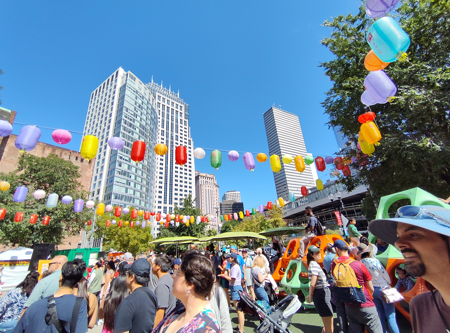 A lively outdoor festival takes place in an urban park with colorful lanterns hanging above a crowd of people against a backdrop of tall city buildings.