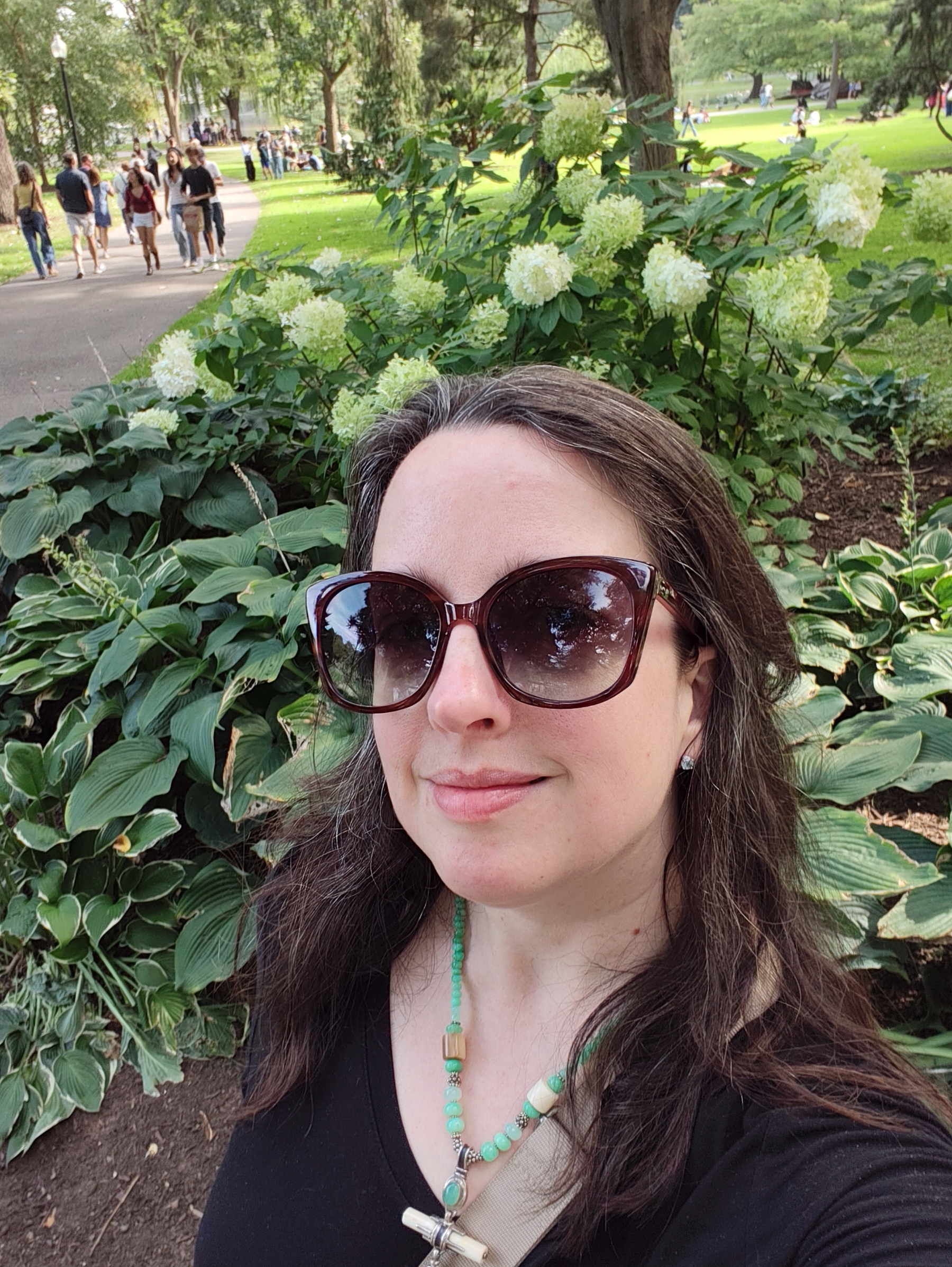 A woman wearing large sunglasses and a beaded necklace takes a selfie in a park with green foliage and blooming white flowers behind her.