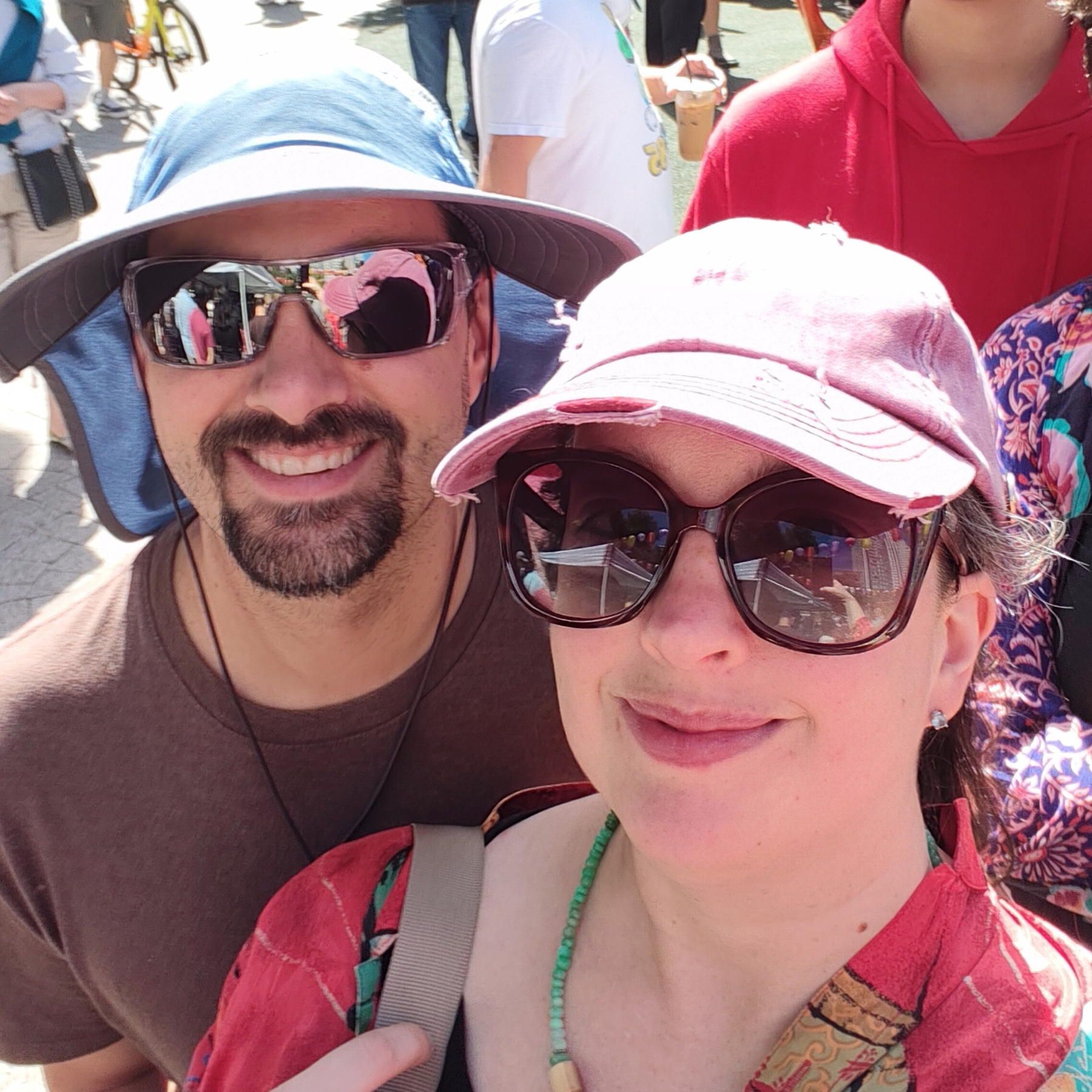 A smiling man and woman wearing sunglasses and hats pose for a selfie outdoors in a sunny setting.