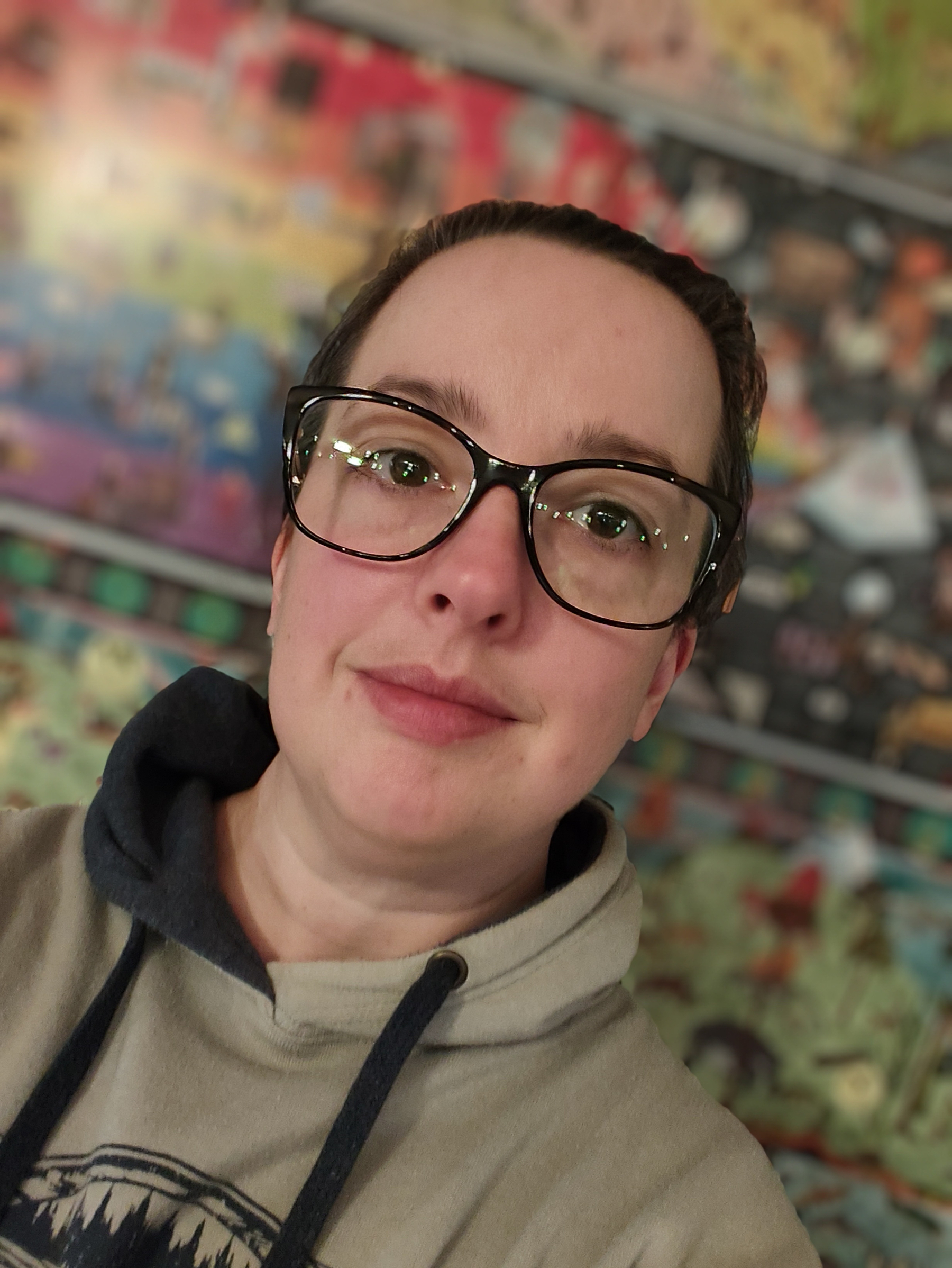 A person wearing large black glasses and a beige hoodie is smiling slightly with a colorful, blurred background behind them.
